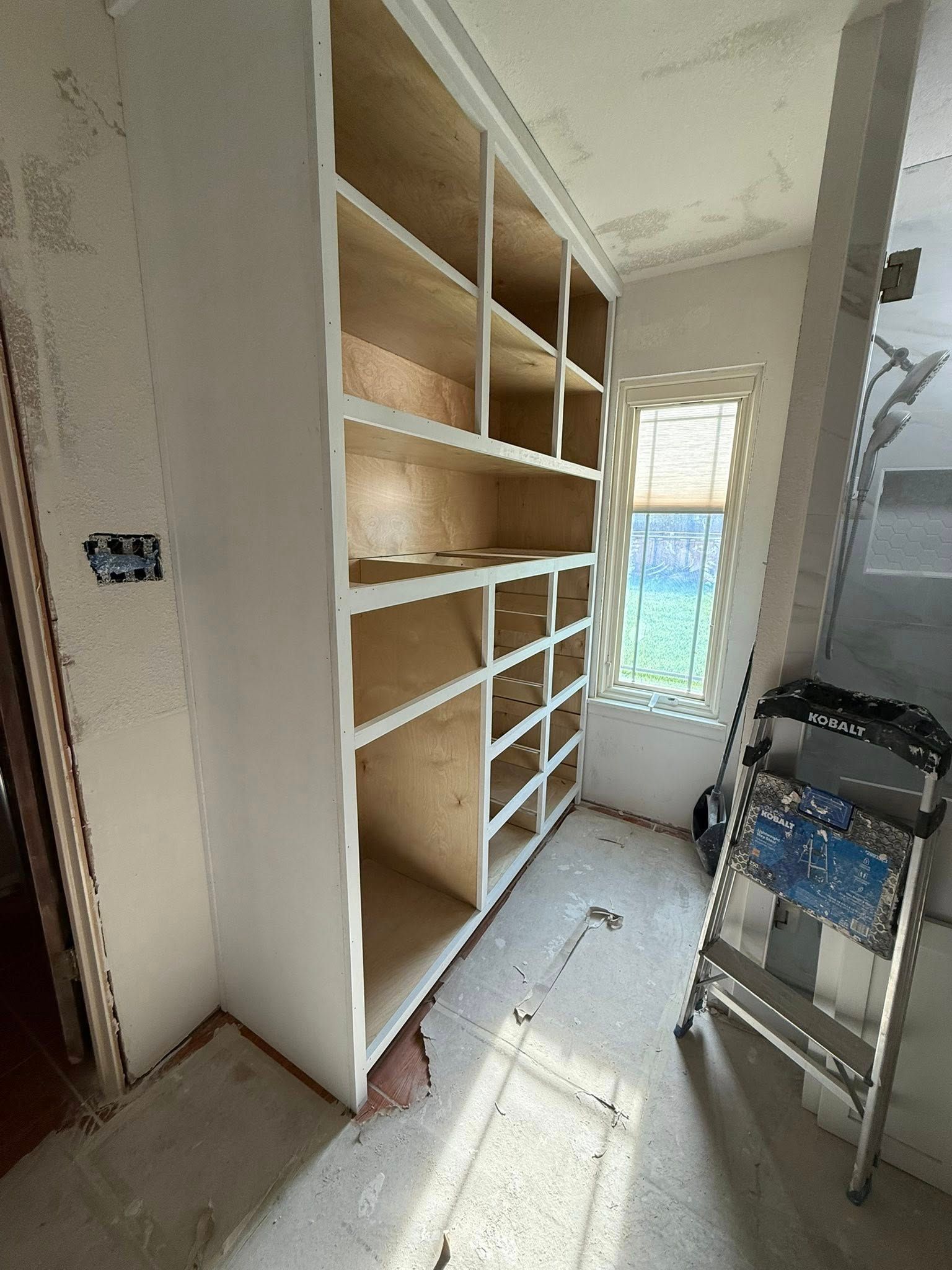 Built-in wooden shelving in a narrow room, with a ladder near a sunlit window.