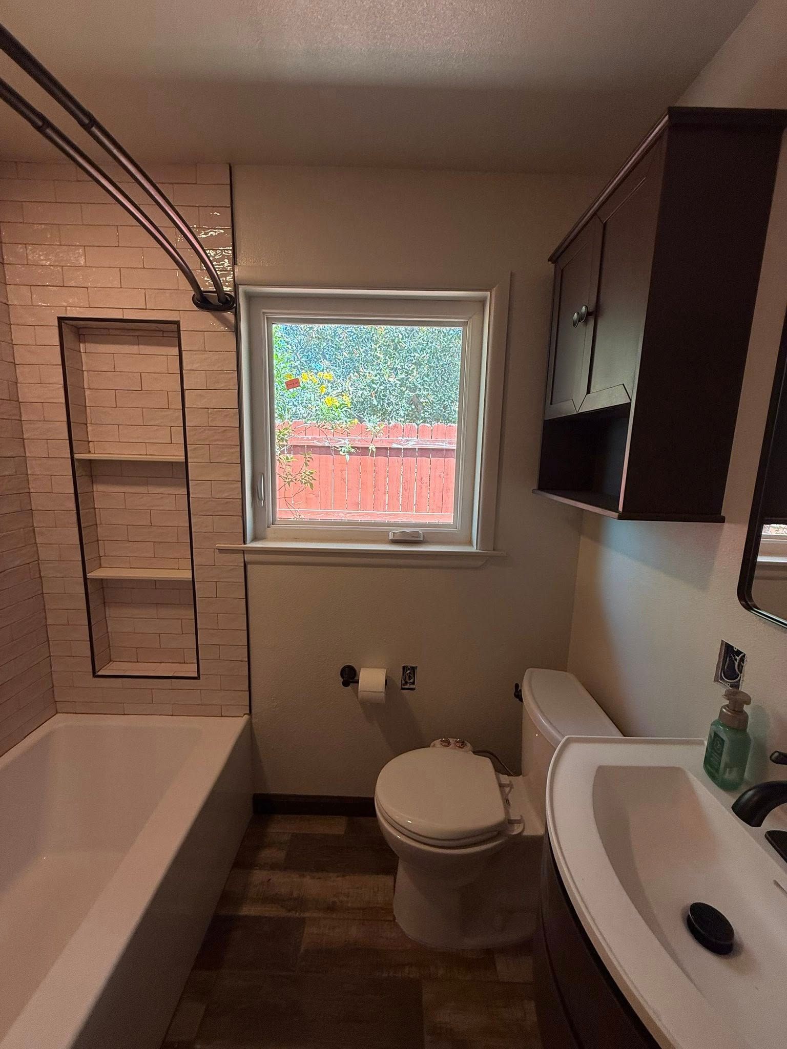 Small bathroom with bathtub, toilet, sink, window, and dark cabinets.