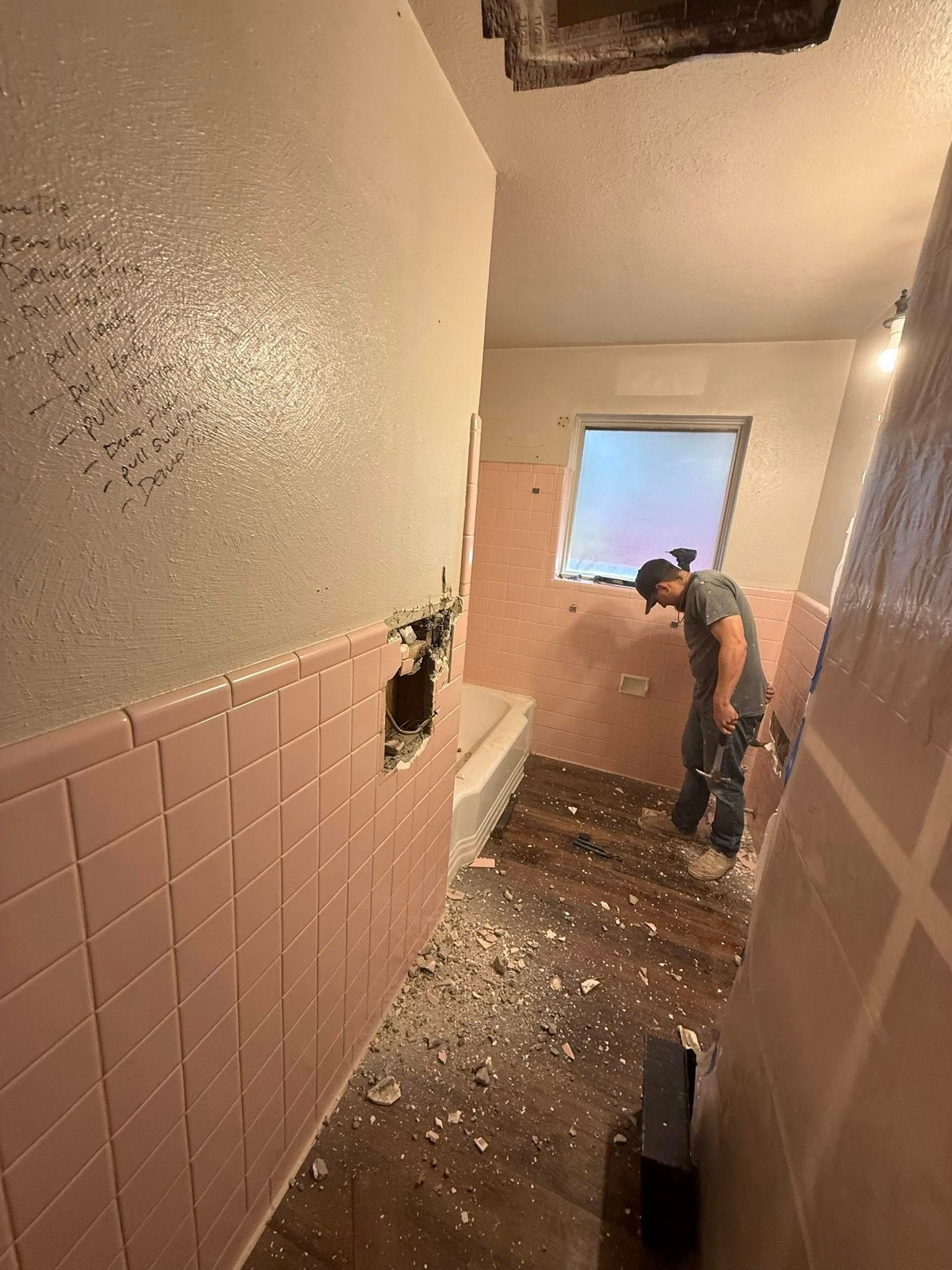 Person cleaning debris in a pink-tiled bathroom hallway with a broken wall and scattered debris.