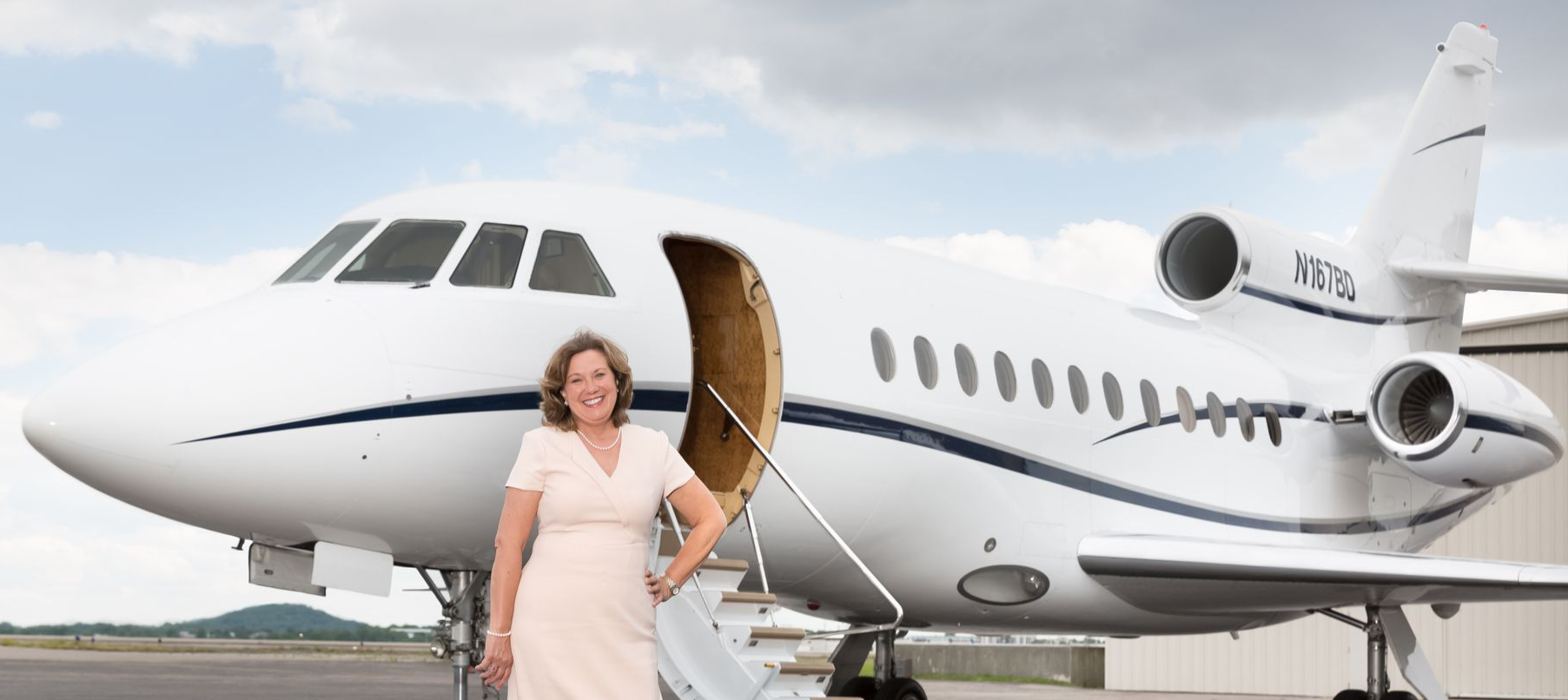 Amanda Rogers at Amjet Aviation; Woman in a peach dress stands beside a private jet on a tarmac, door open.
