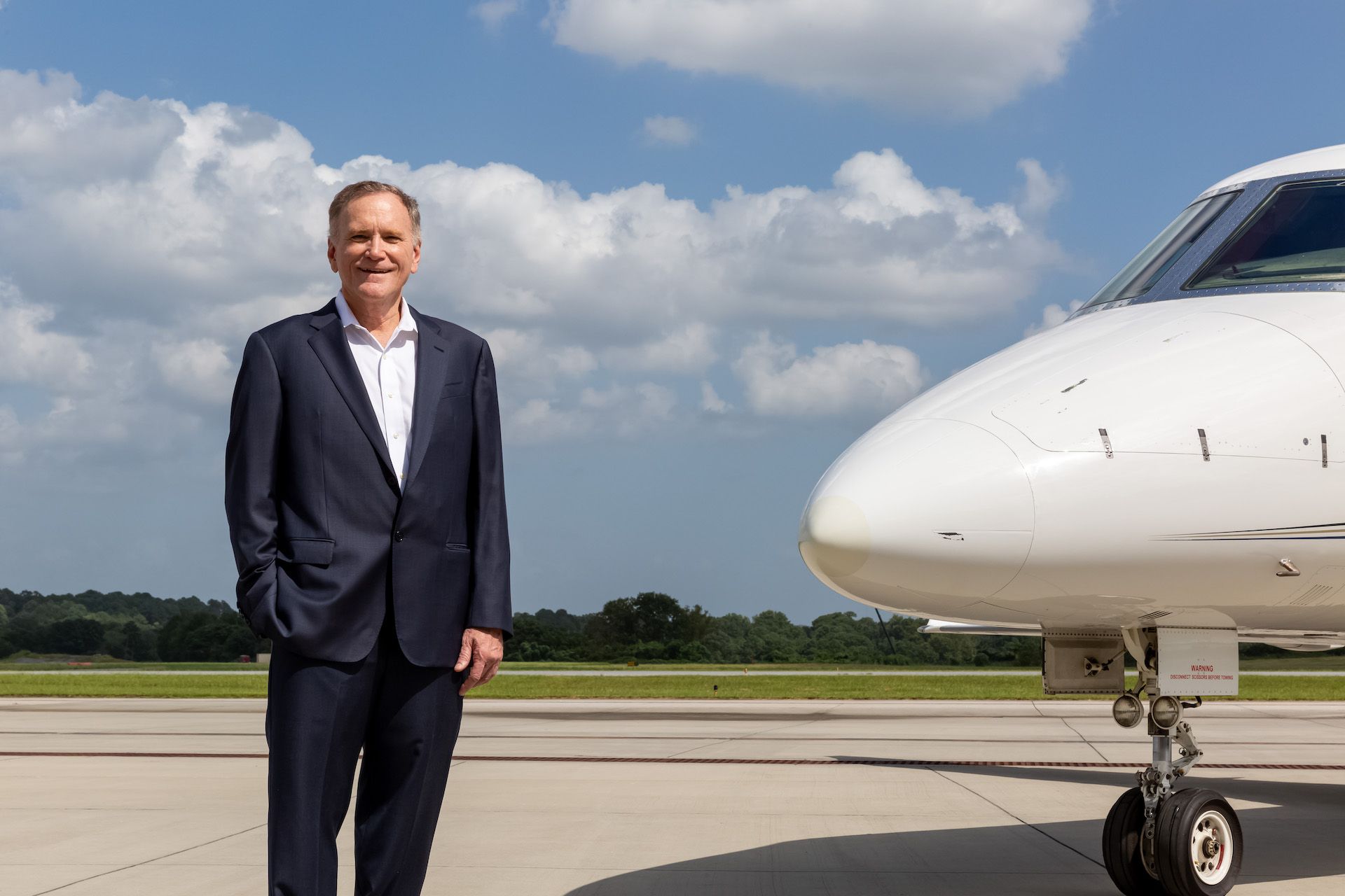 Thomas Rogers at Amjet Aviation;
Man in a suit stands beside a private jet on a sunny tarmac with a blue sky.