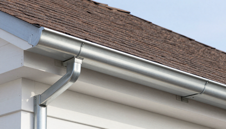 Close-up of a silver metal gutter along a brown shingled roof edge on a white house
