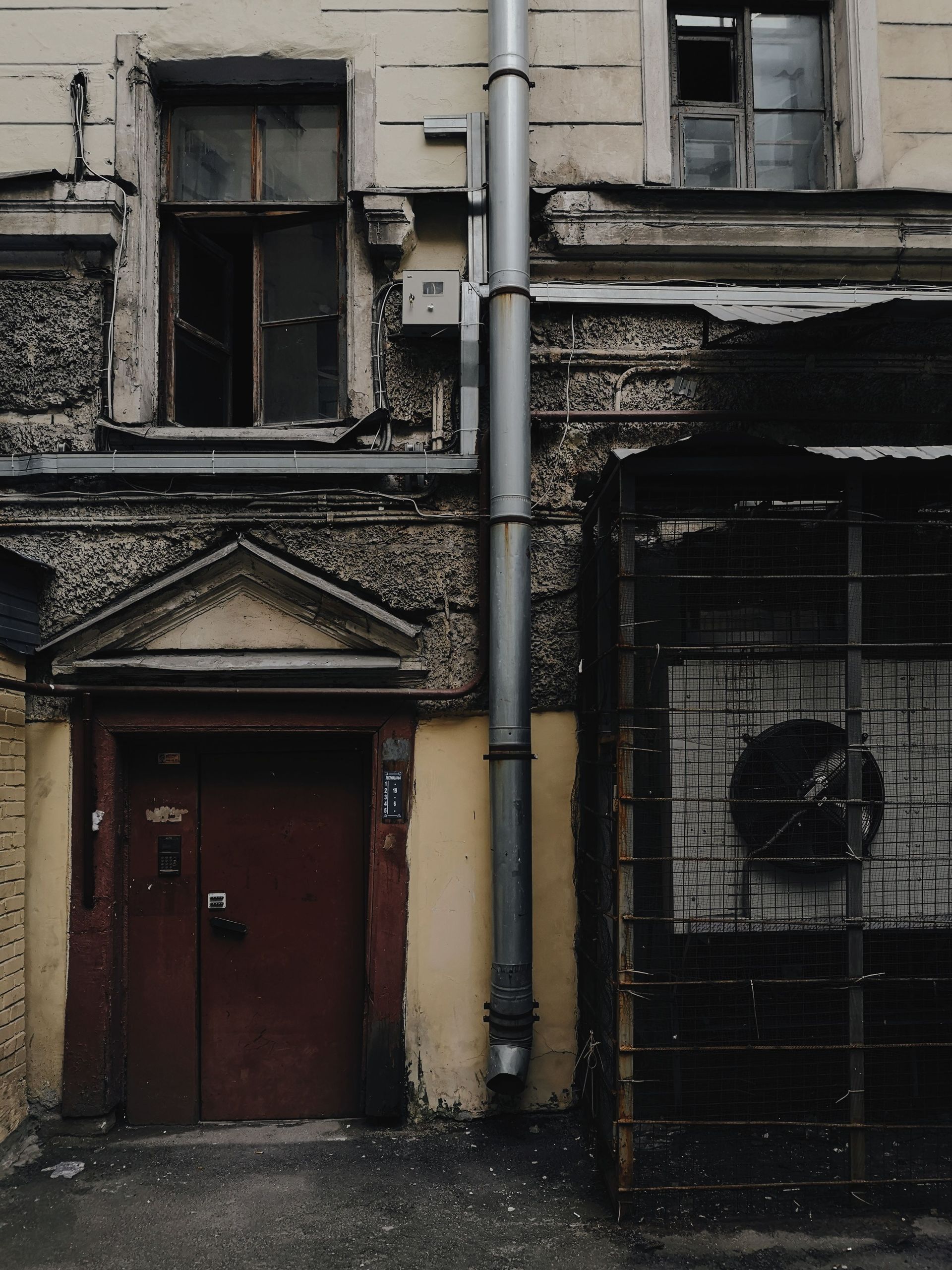 Old weathered building façade with a red door, barred window, and vertical drainpipes.