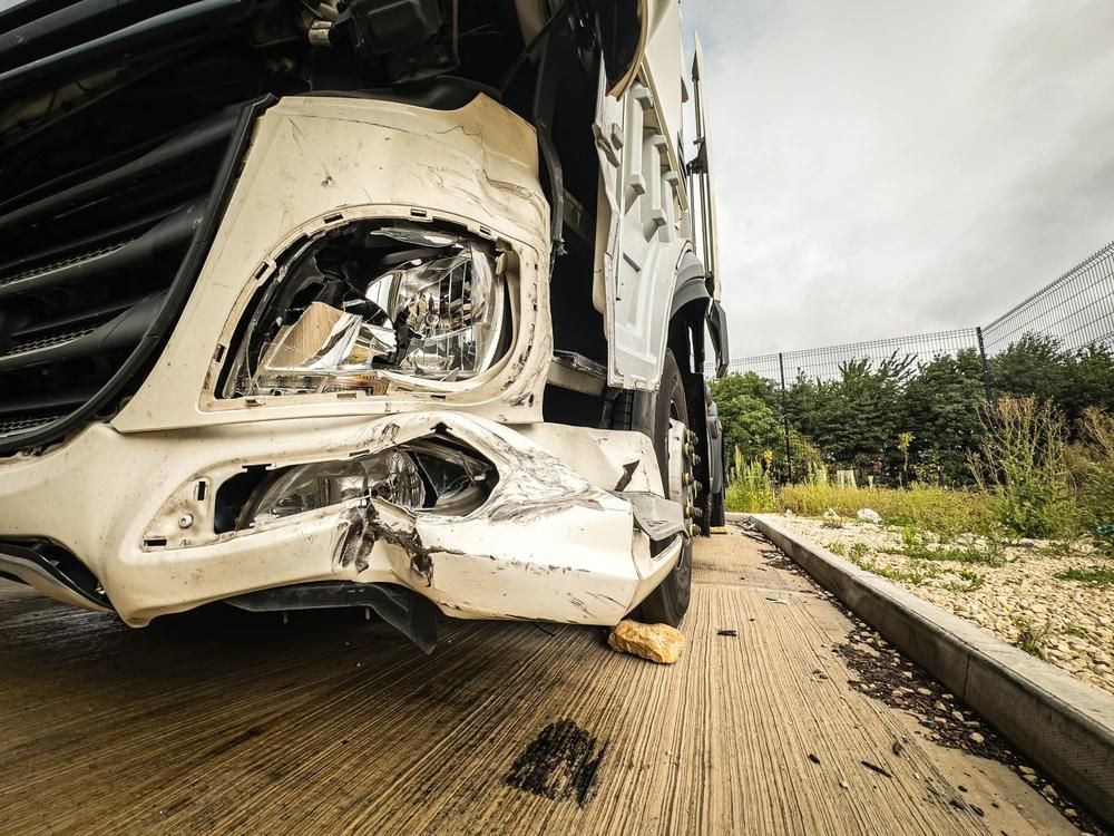 Damaged white truck's front end, visible headlight, bent bumper, on roadside, damaged by a crash.