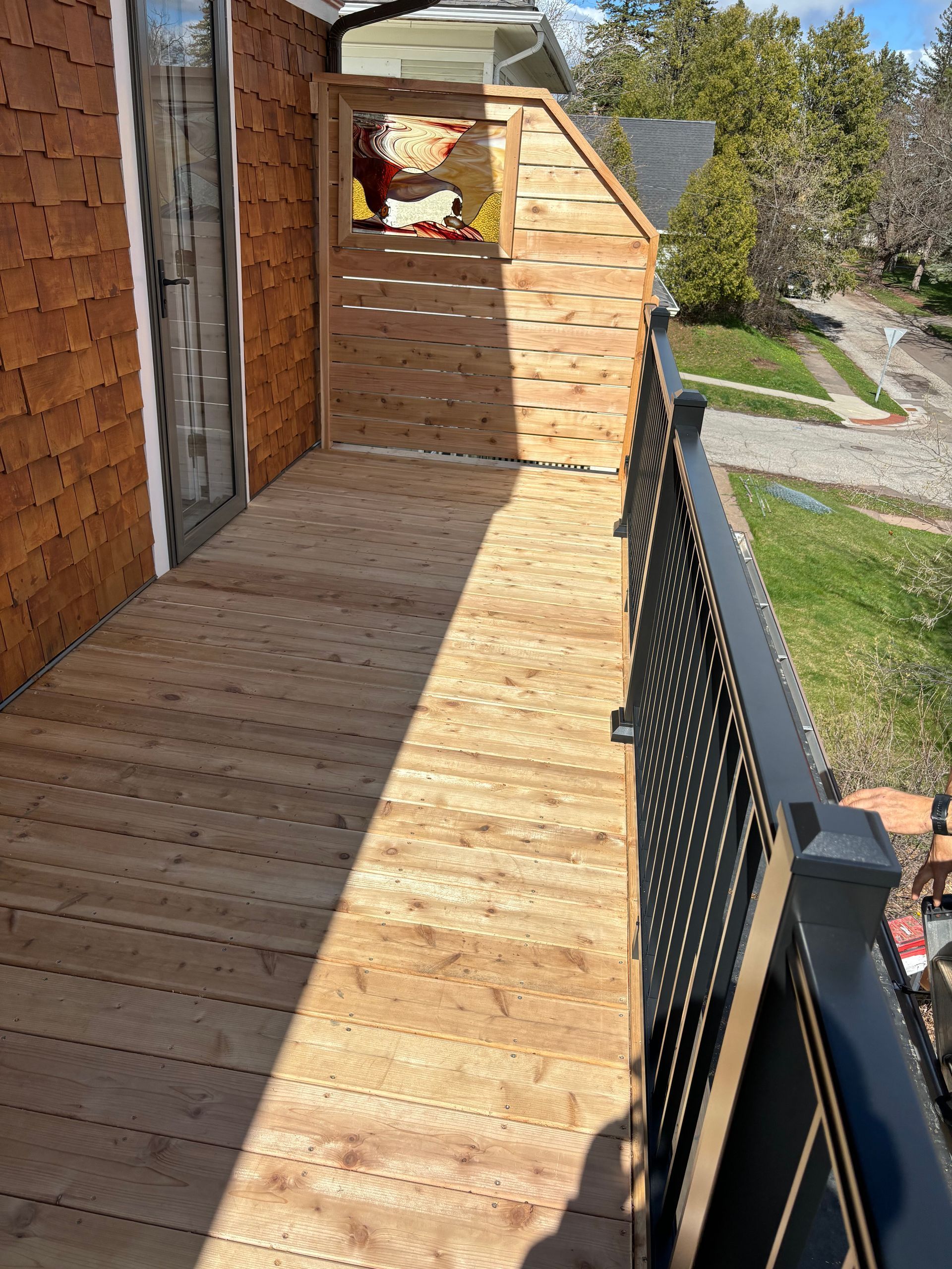 Wooden balcony with black railing, wooden deck, wooden privacy screen, door to home on the left.