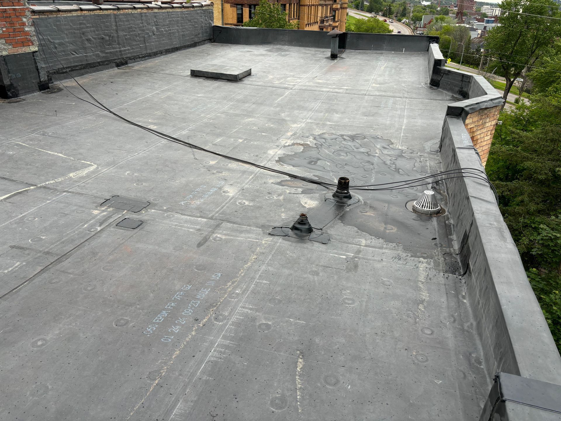 Flat, gray tar roof with various black attachments and water stains, bordered by a brick wall and green trees.