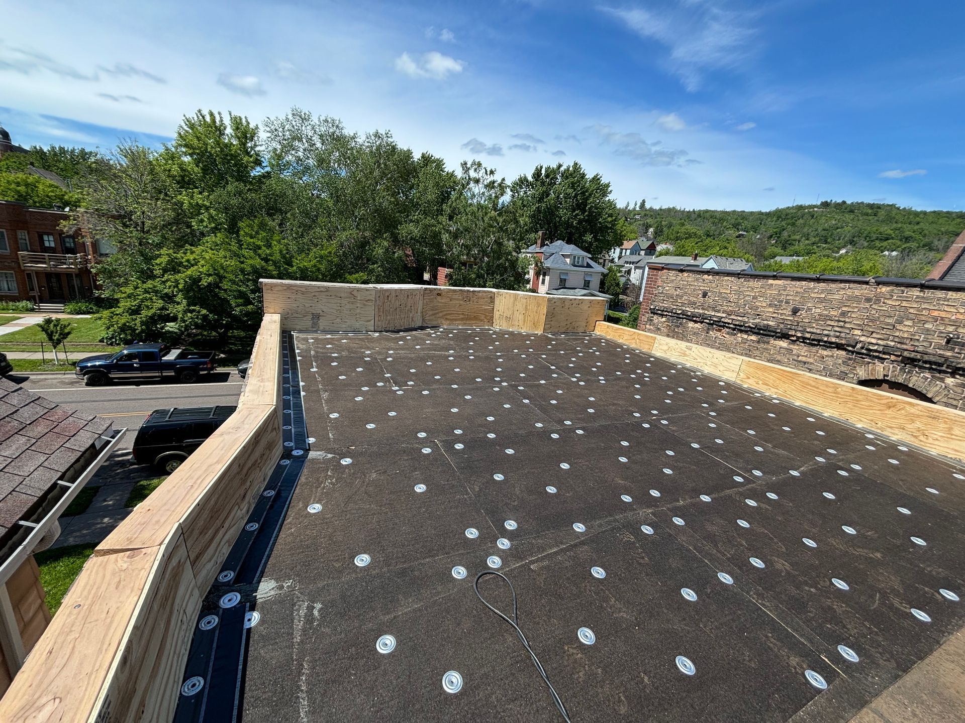 Rooftop with black roofing material, wooden trim, and numerous white circular fasteners, under a blue sky.