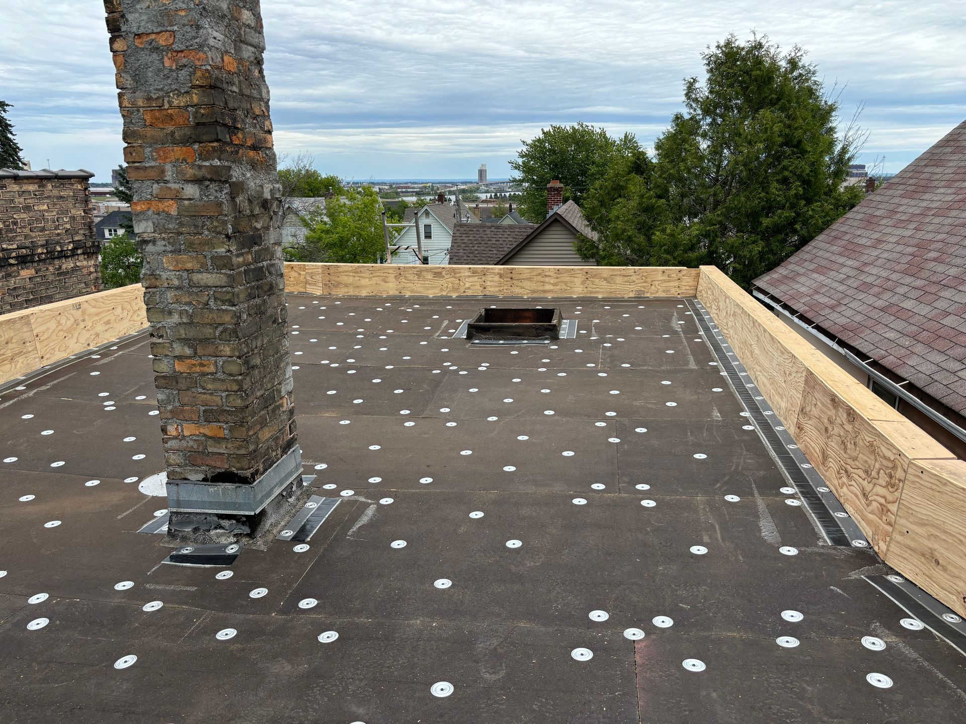 Flat roof with brick chimney, black surface, white fasteners. Wooden perimeter, cityscape in background.