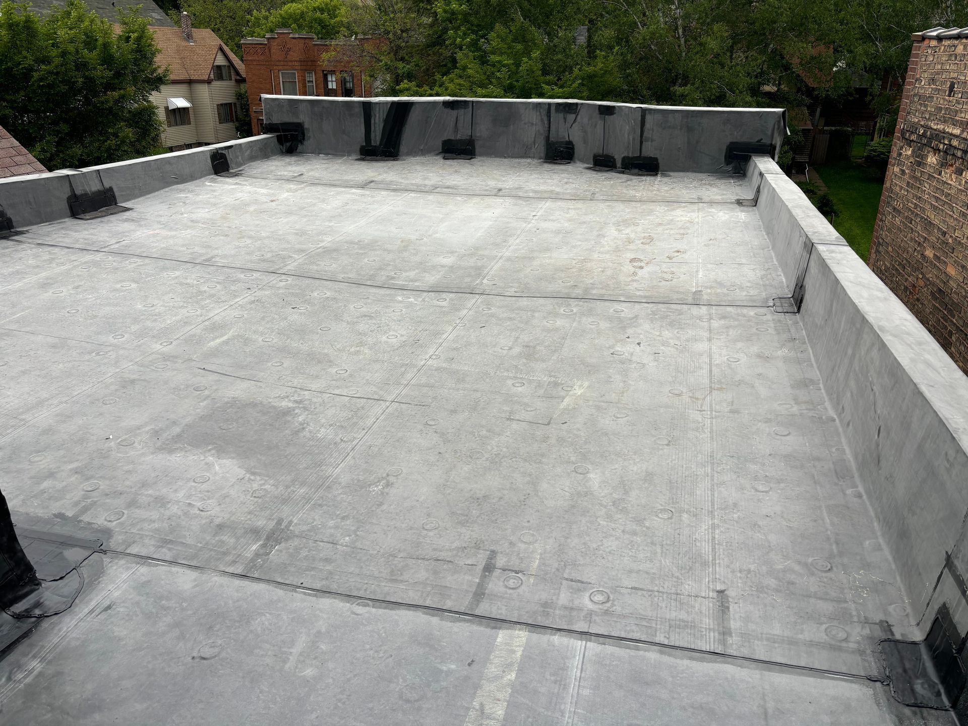 Flat, gray rooftop with concrete edging. Black sealants and small buildings visible in background.