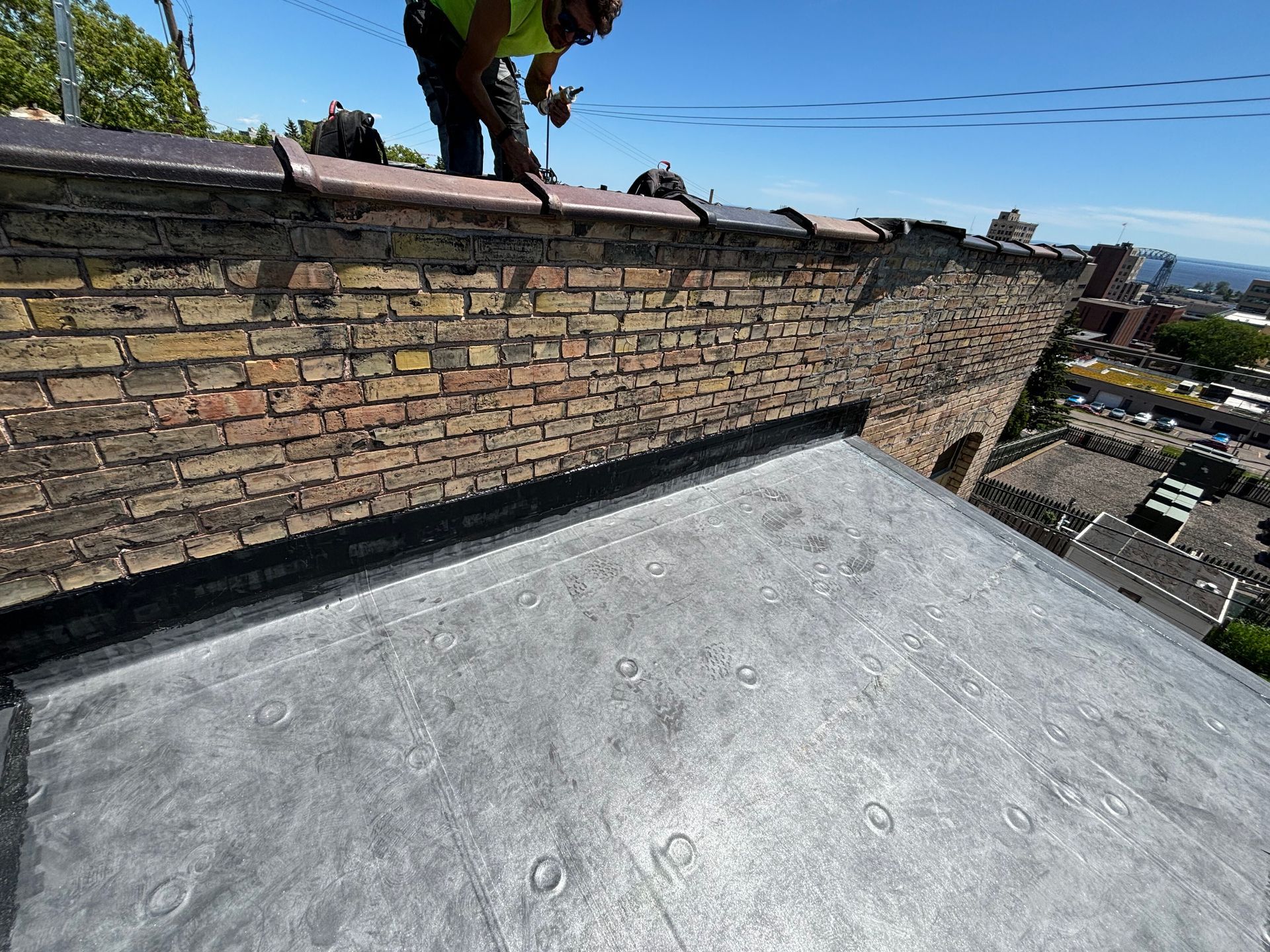 Roofer in safety gear works on a flat, black roof near a brick wall on a sunny day. City in the background.