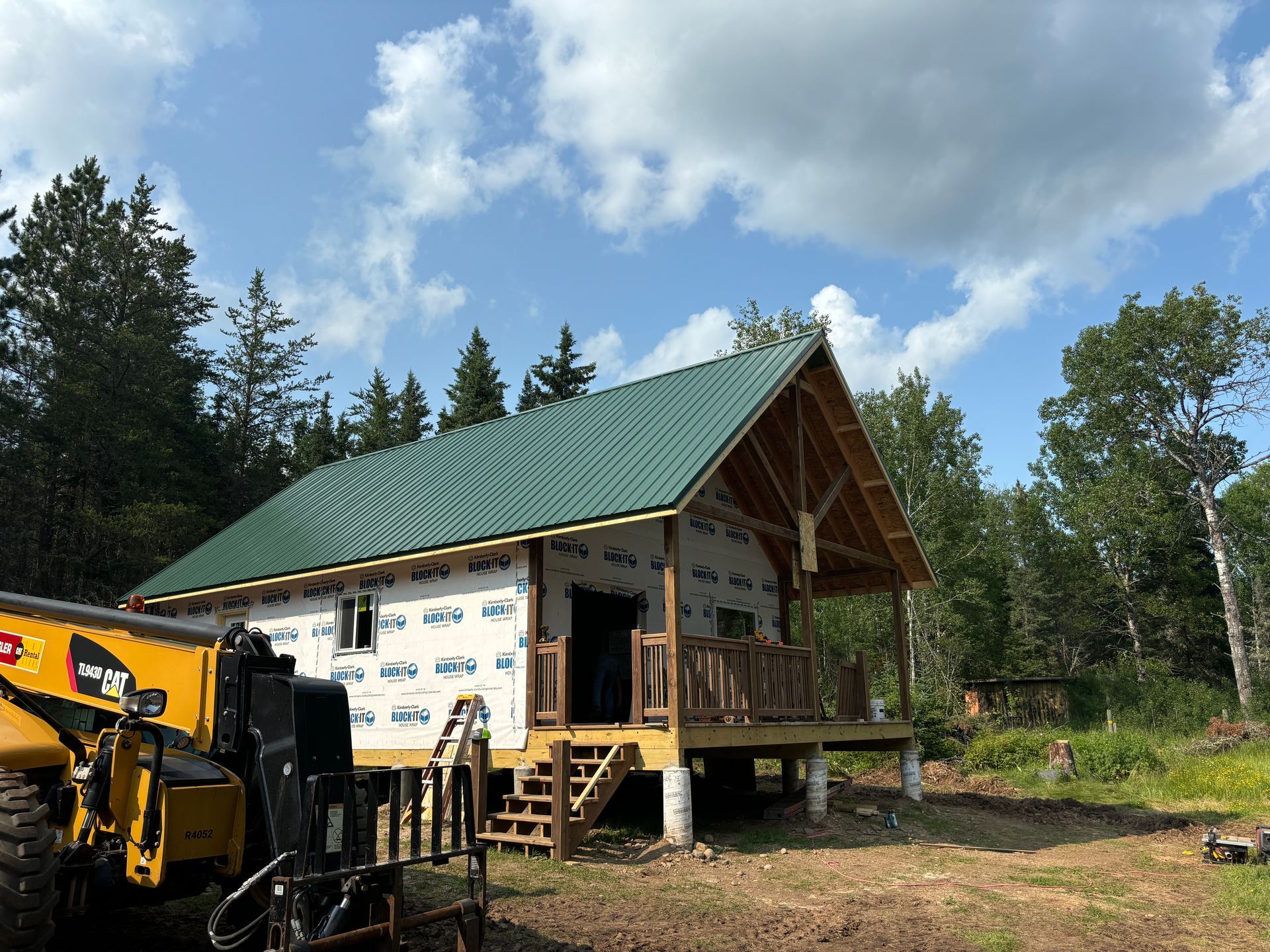 Cabin under construction with green metal roof, wrap, porch, and stairs in a wooded area.