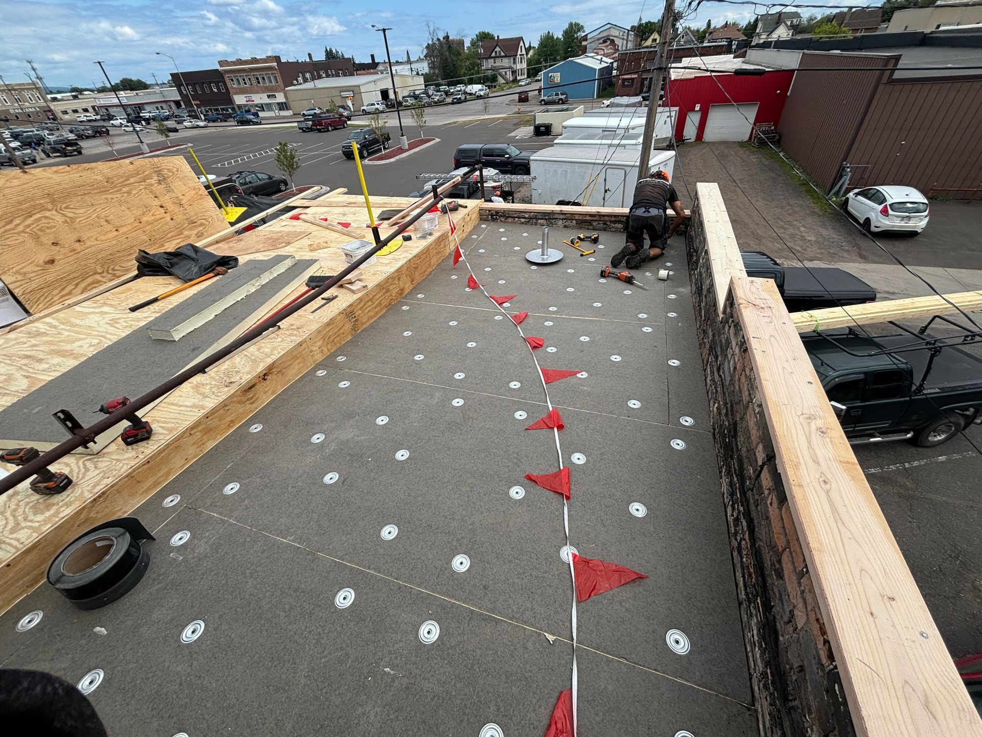 Roofing work in progress on a flat roof. A person works near red flags. Buildings and vehicles are in the background.