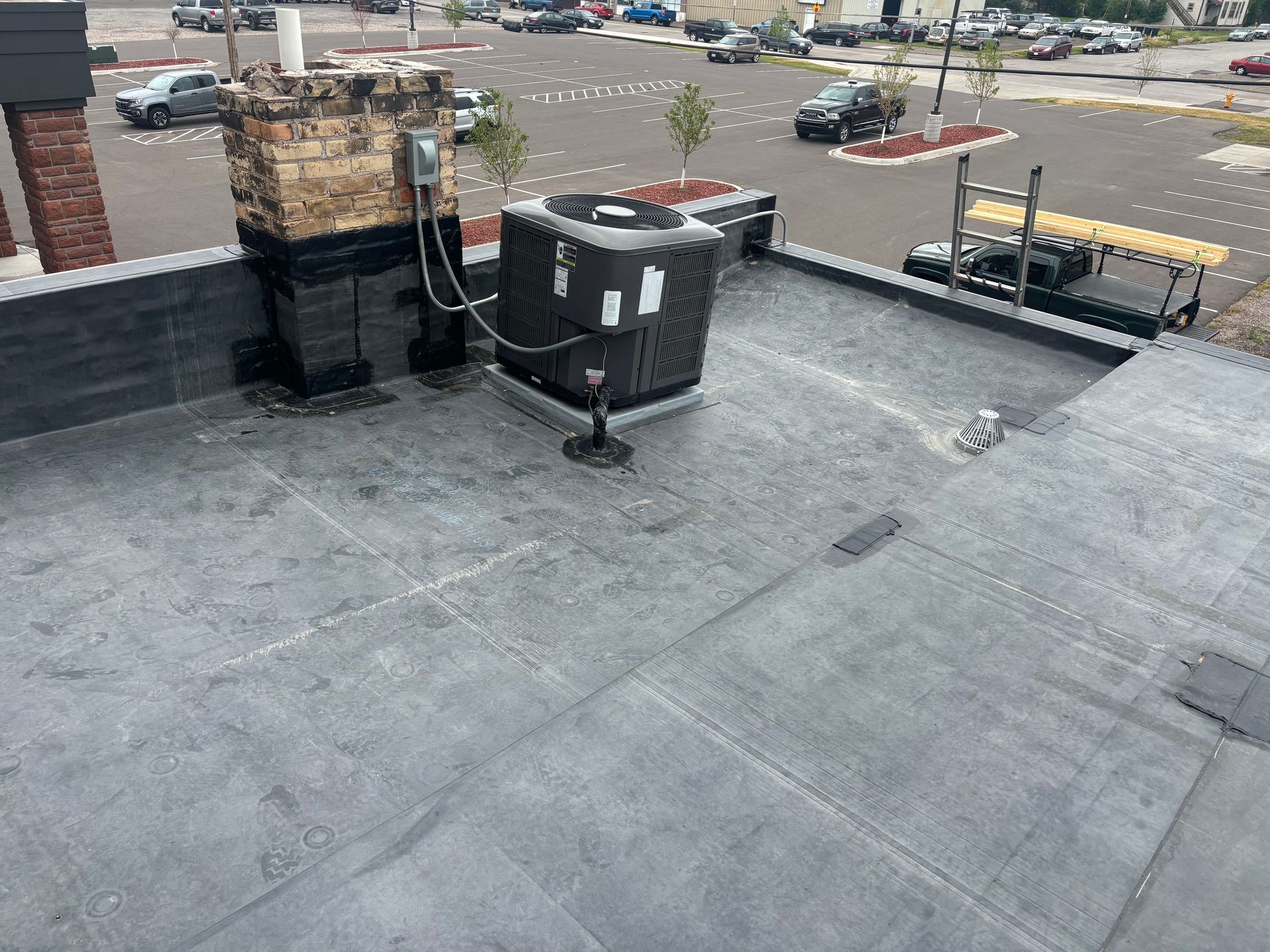 Black commercial rooftop with HVAC unit, brick chimney, and parking lot view.