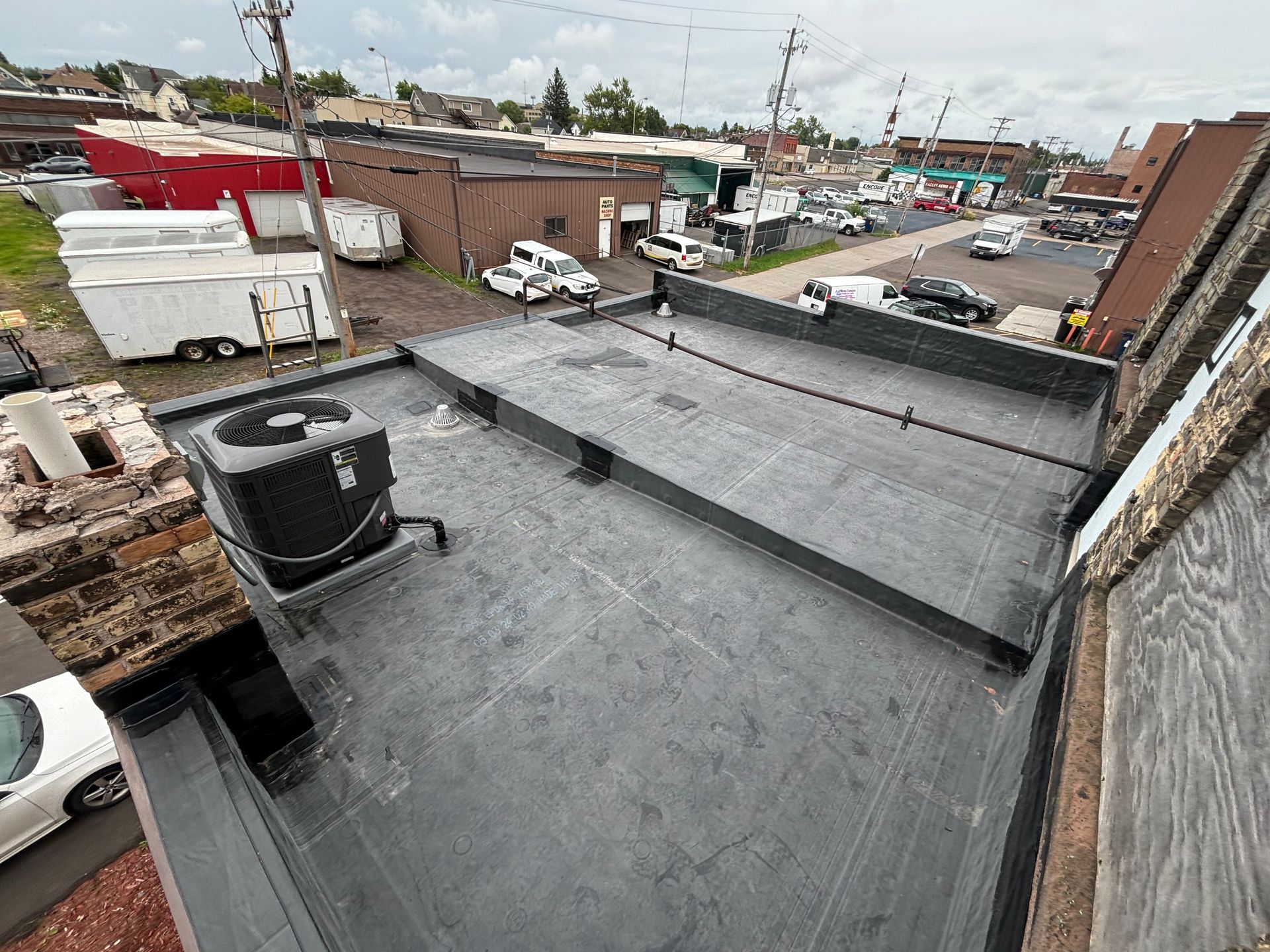 Overhead view of a flat, black roof with air conditioning unit; buildings and vehicles in the background.