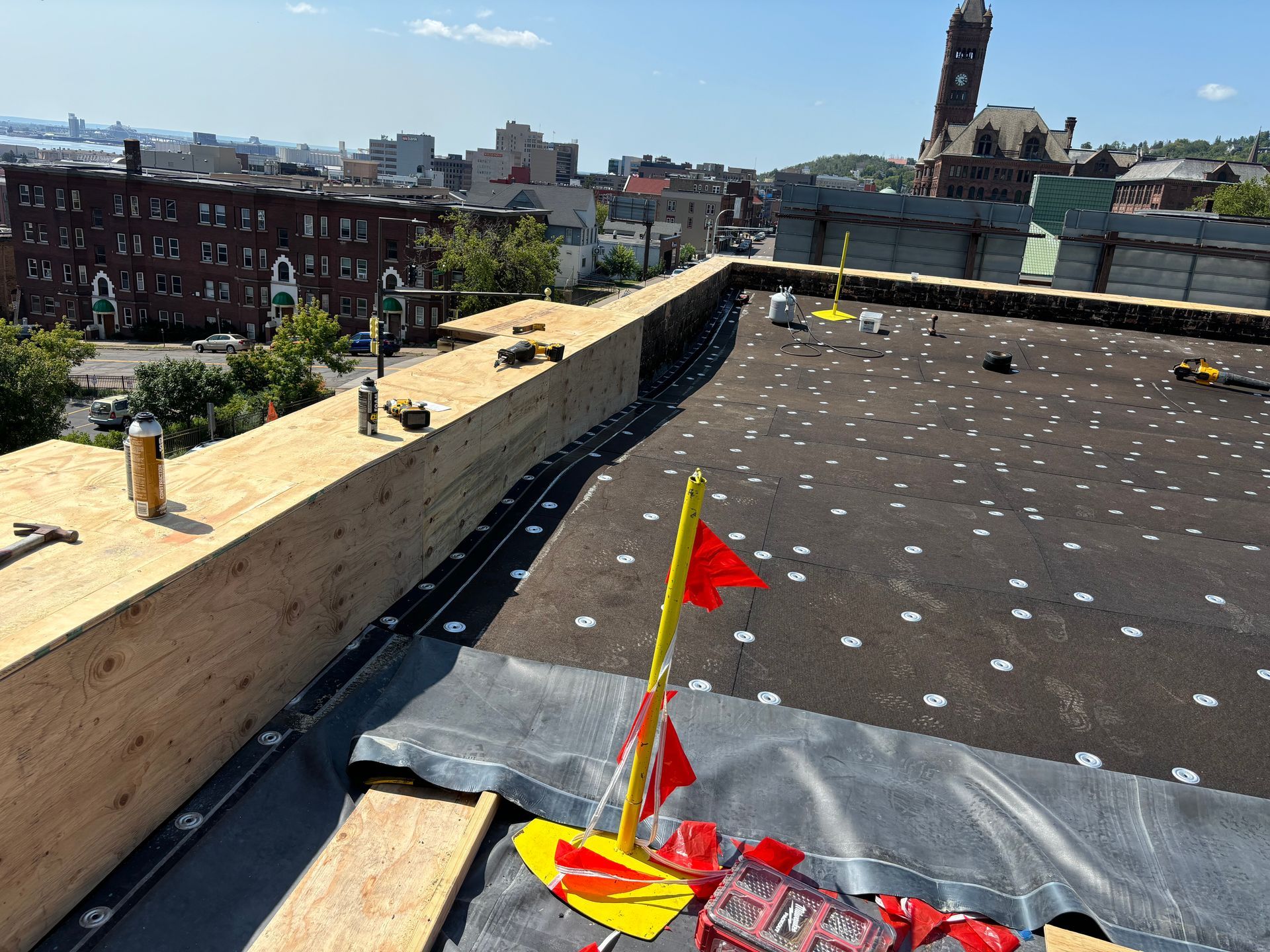 Rooftop construction with wooden barrier and dark roofing material. City skyline visible in background.