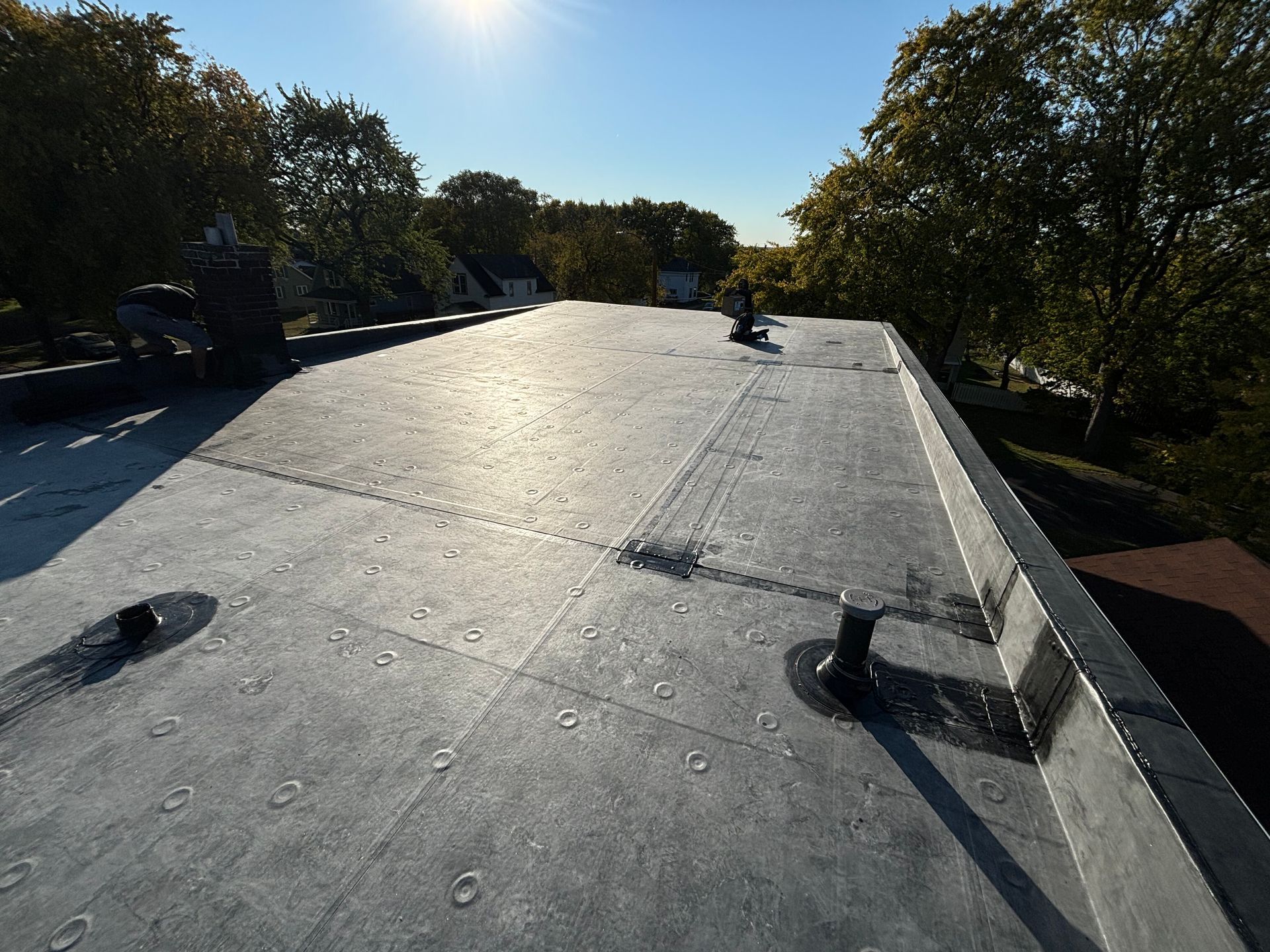 Flat, black roof with several plumbing vents; two people near chimney; sunny day.