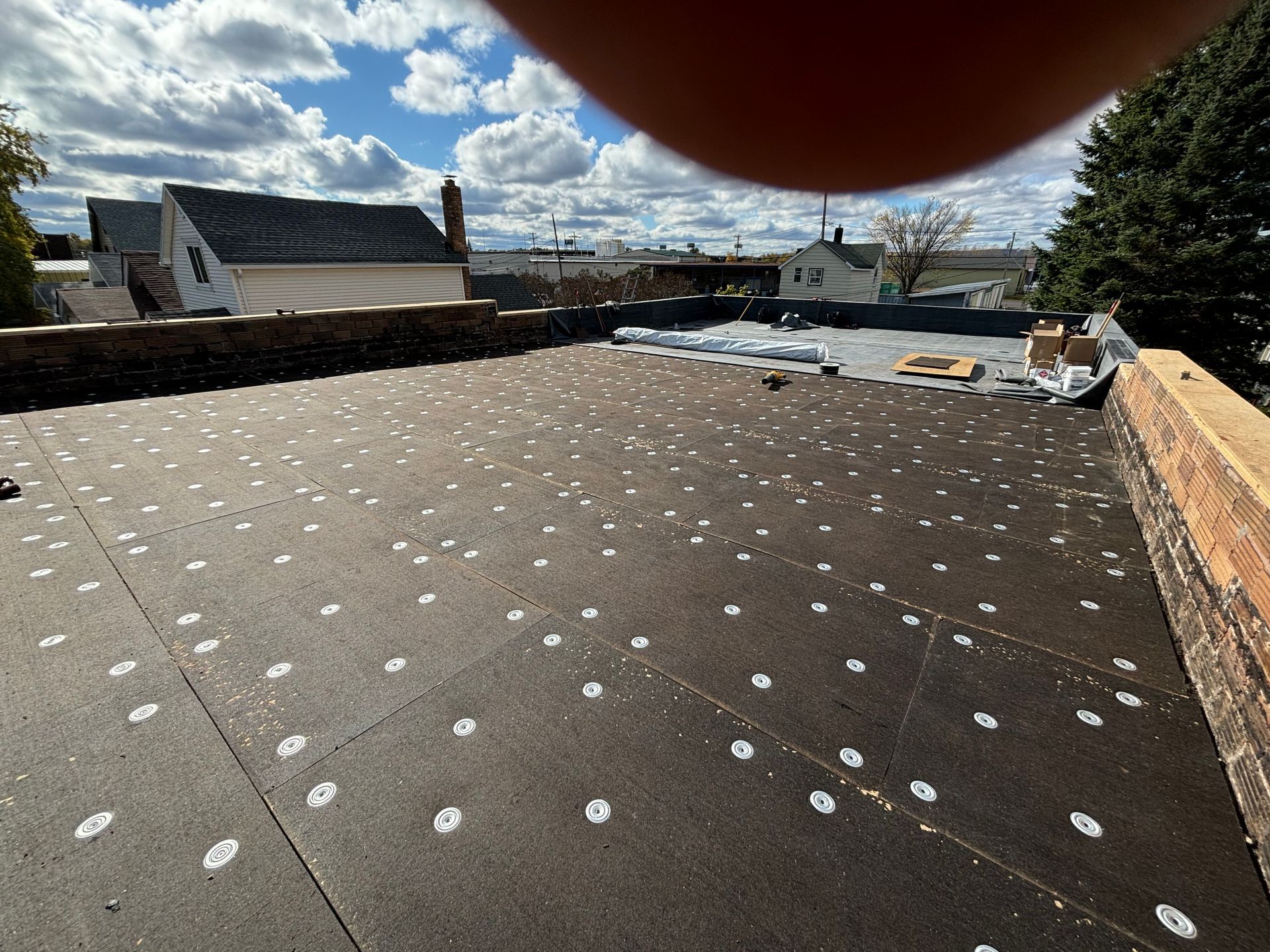 A flat, black rooftop with white fasteners, under a cloudy sky. Brick wall on the right.