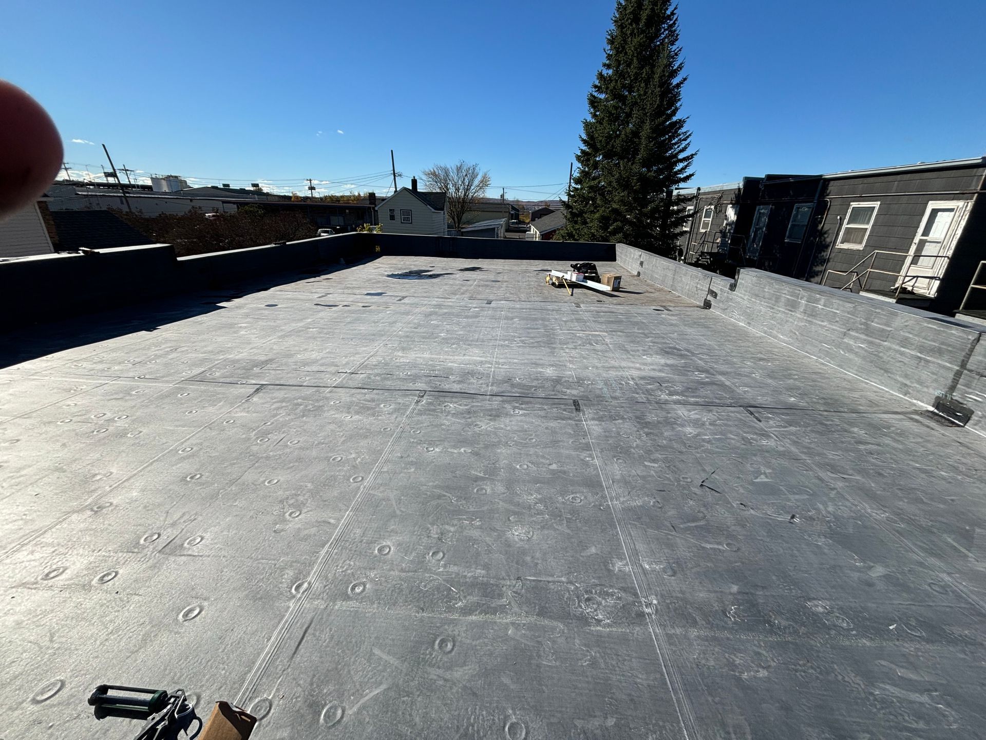 Flat, dark rooftop on a sunny day. A building is under construction, and a tall evergreen tree is visible in the background.
