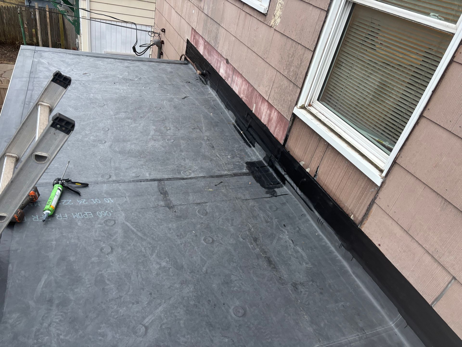 Flat roof with ladder, sealant, and building exterior. Black roofing material with caulk applied at the wall's edge.
