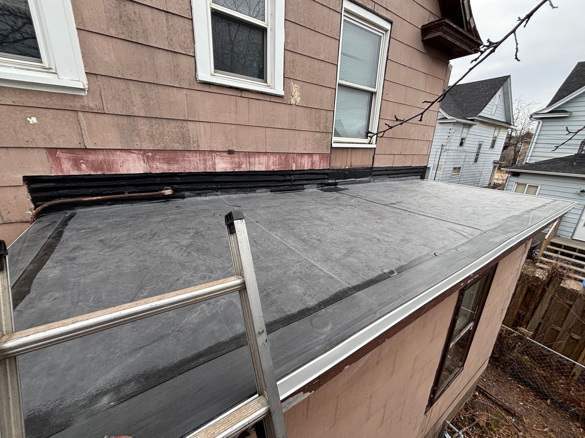 A black flat roof with a ladder leaning against it. An older building in the background.