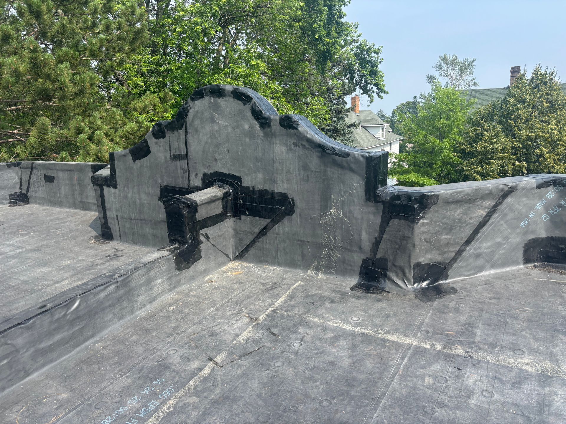 Black flat roof with sealed seams and parapet against a backdrop of trees and houses.