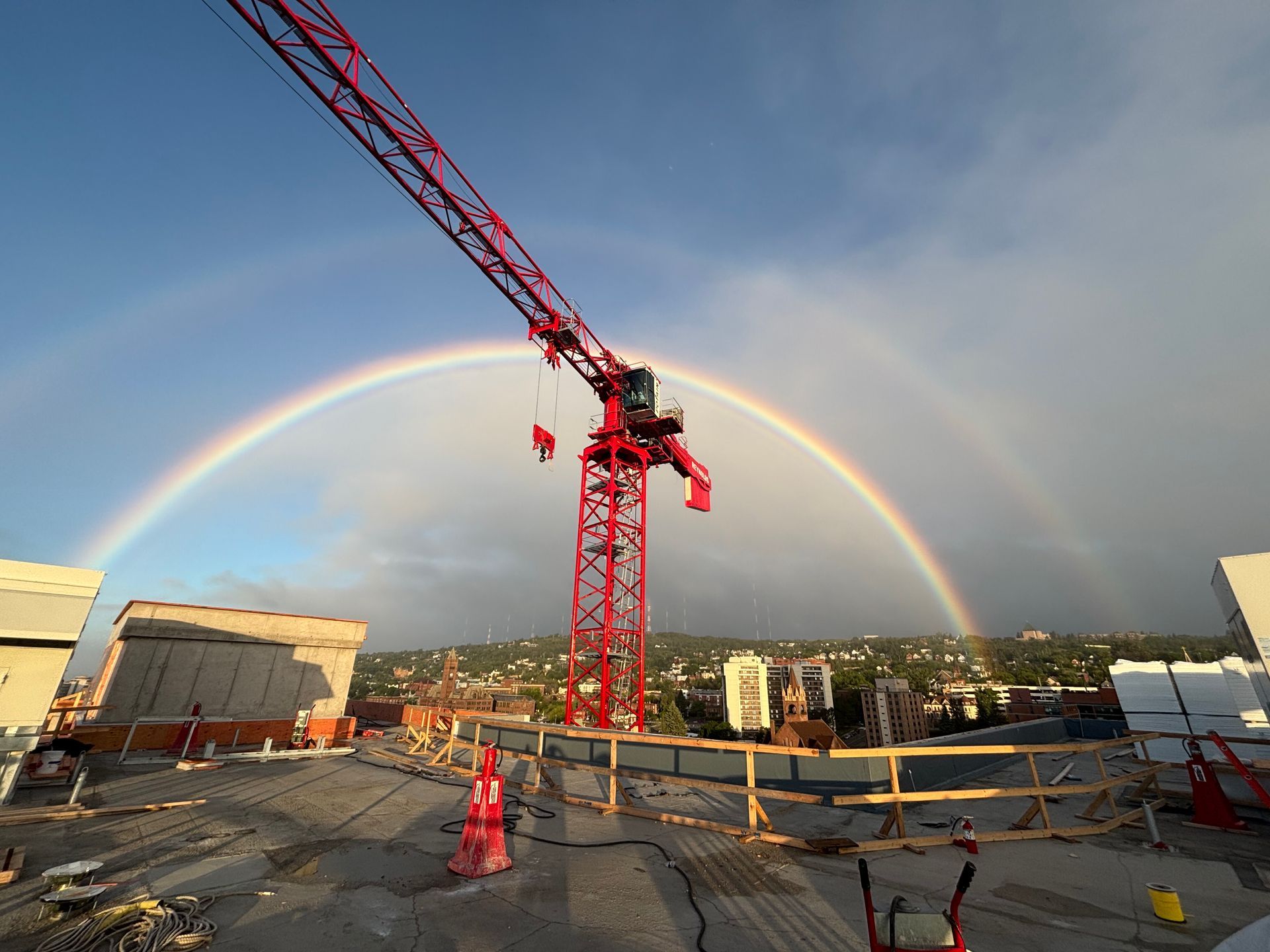 Construction site with red crane against a double rainbow arching over a city.