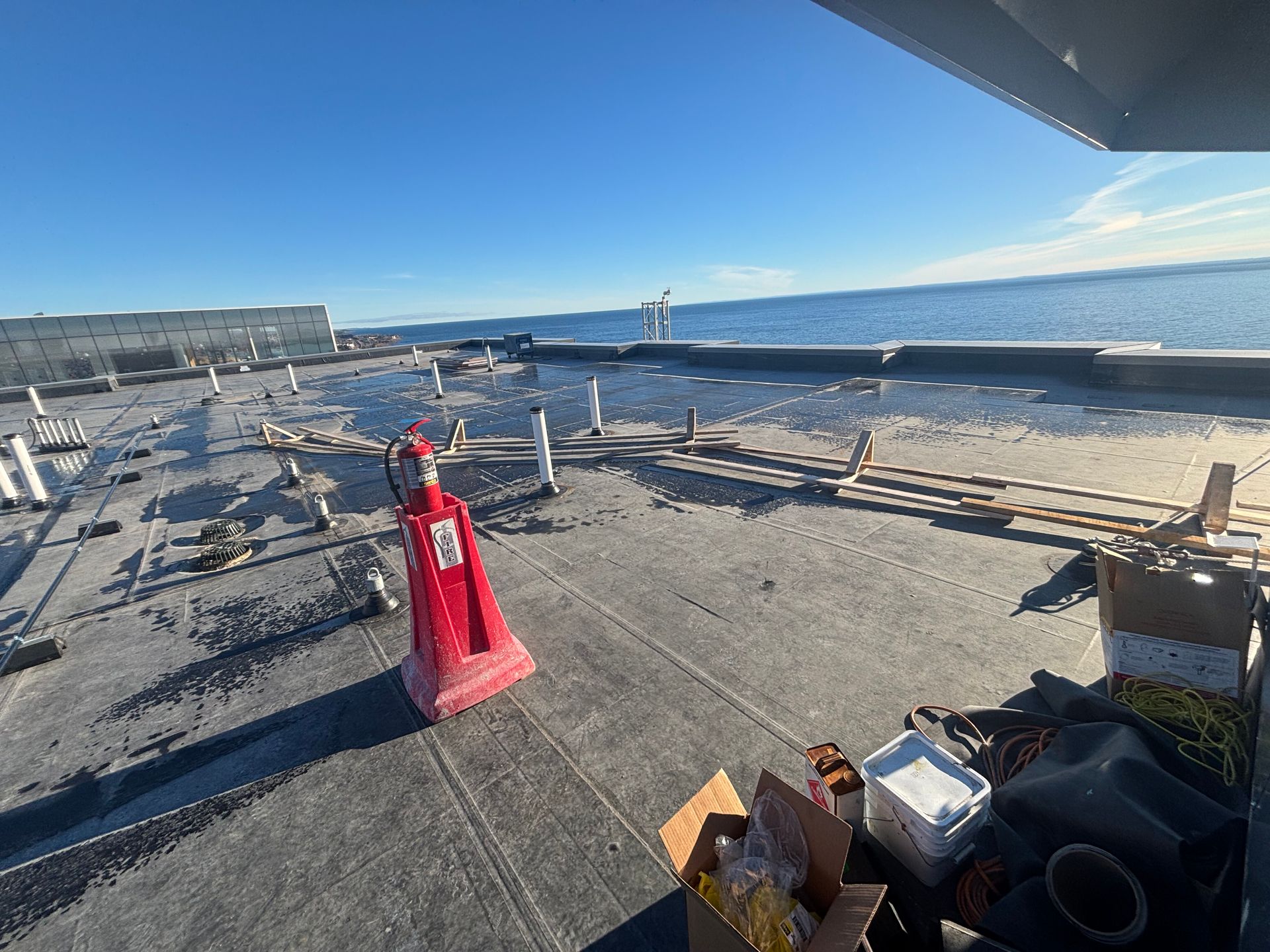 Rooftop with construction materials, pipes, and a bright blue sky overlooking water. A red safety barricade is visible.