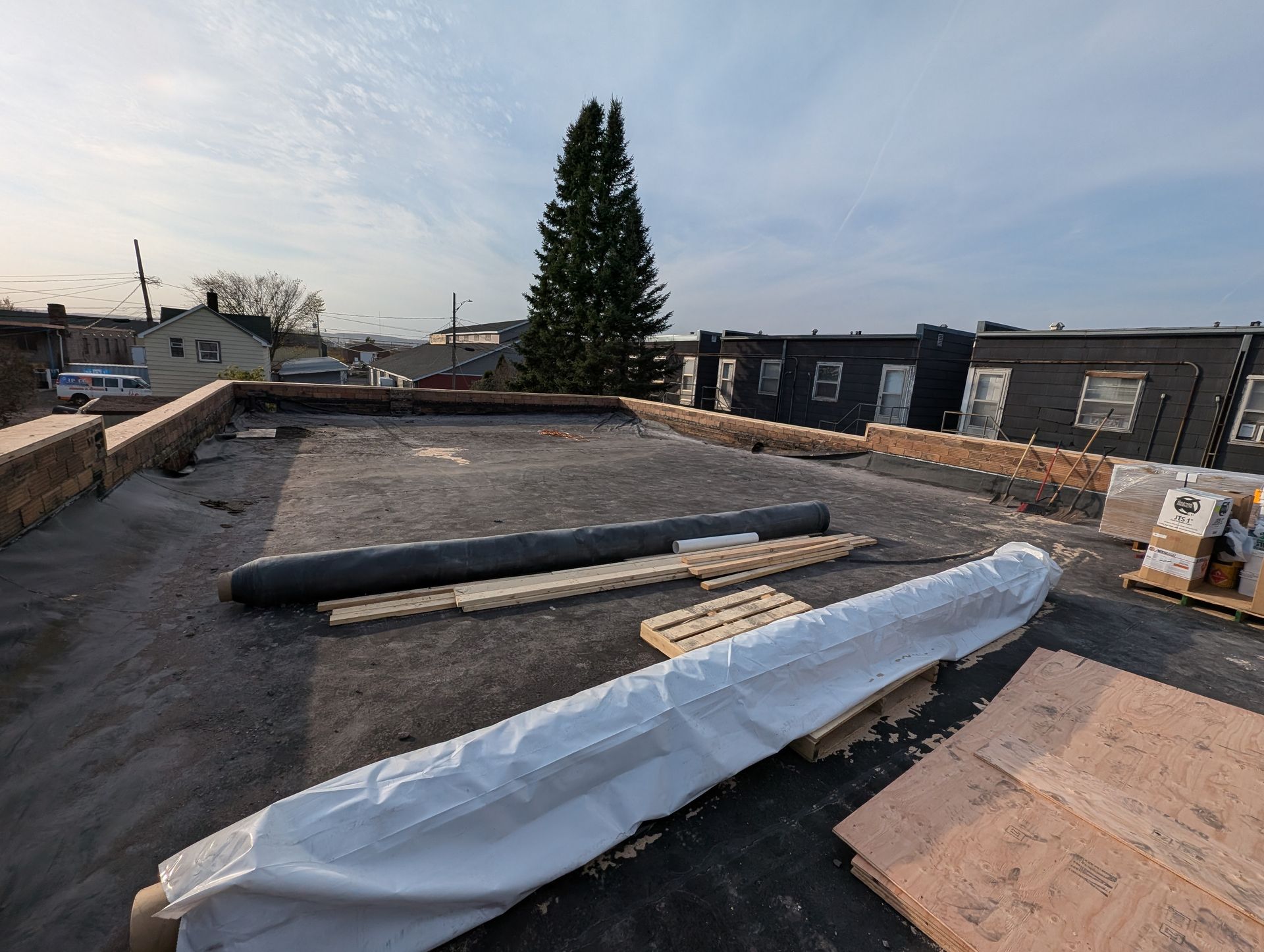 Rooftop with construction materials: rolls, wooden planks, and partially covered surface. Large evergreen tree in distance.