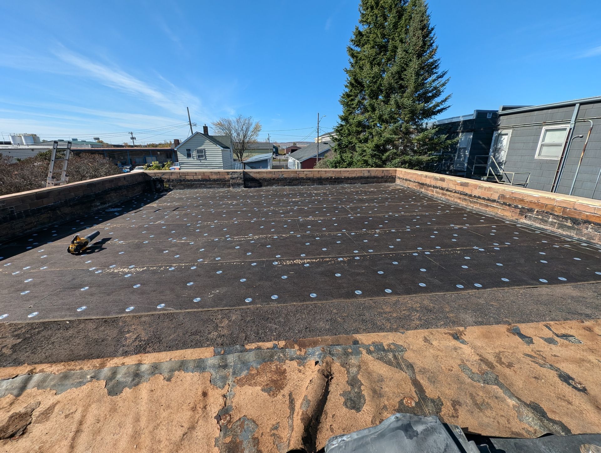 Flat rooftop with black roofing material, scattered debris, and surrounding buildings under a blue sky.