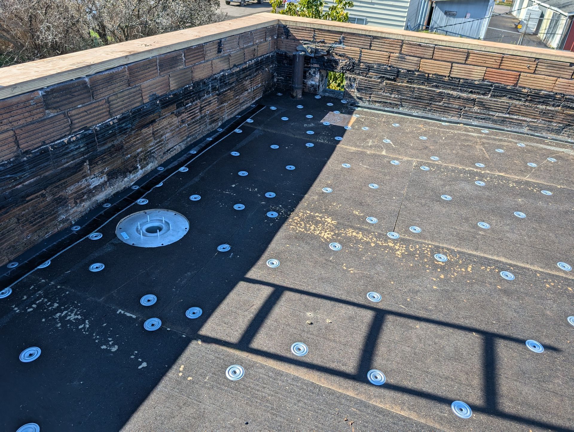 Flat tar roof with circular fasteners and drain. Brick wall surrounds the edge.