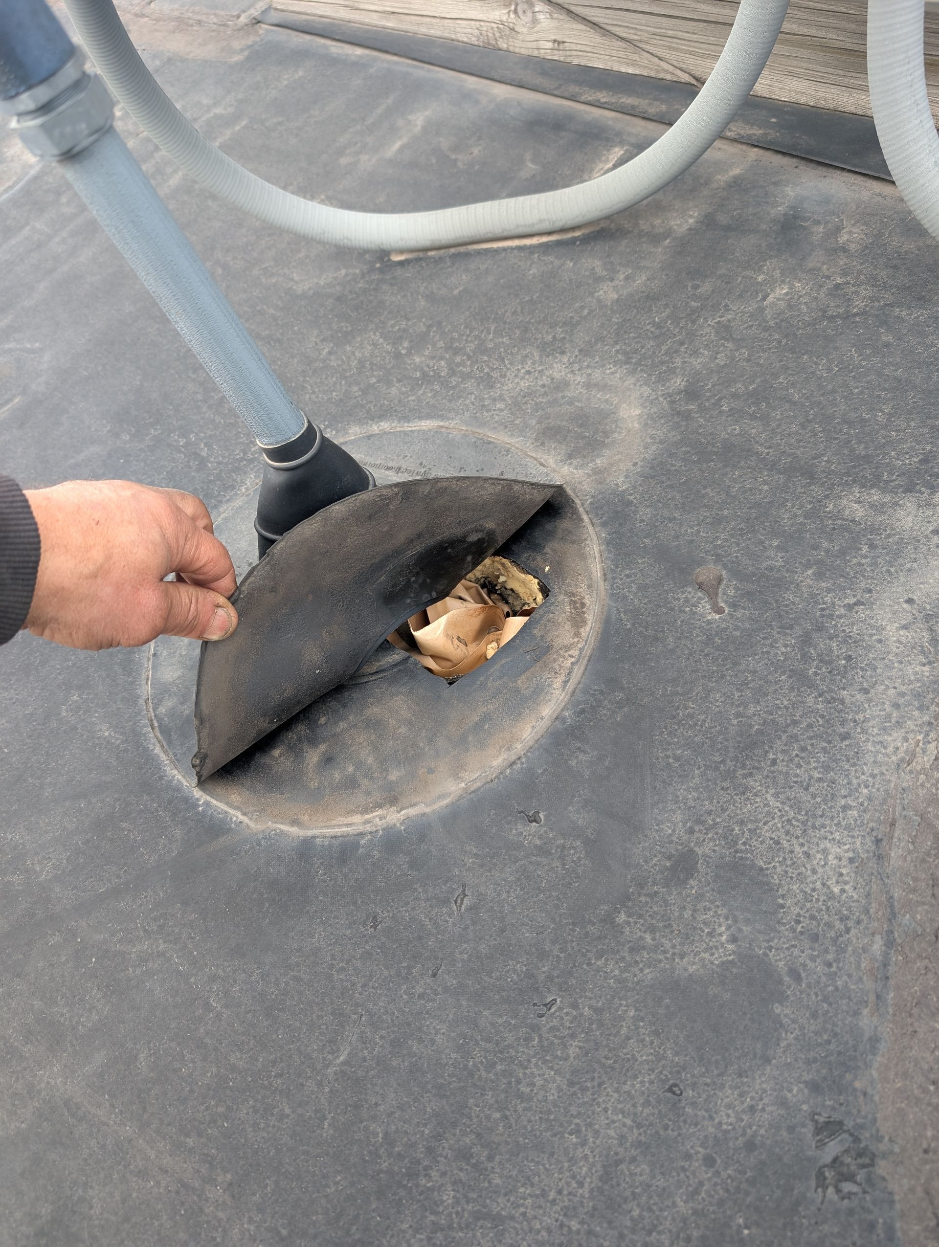 Person lifting a rubber flap covering a drain opening. Gray surface with a hose.