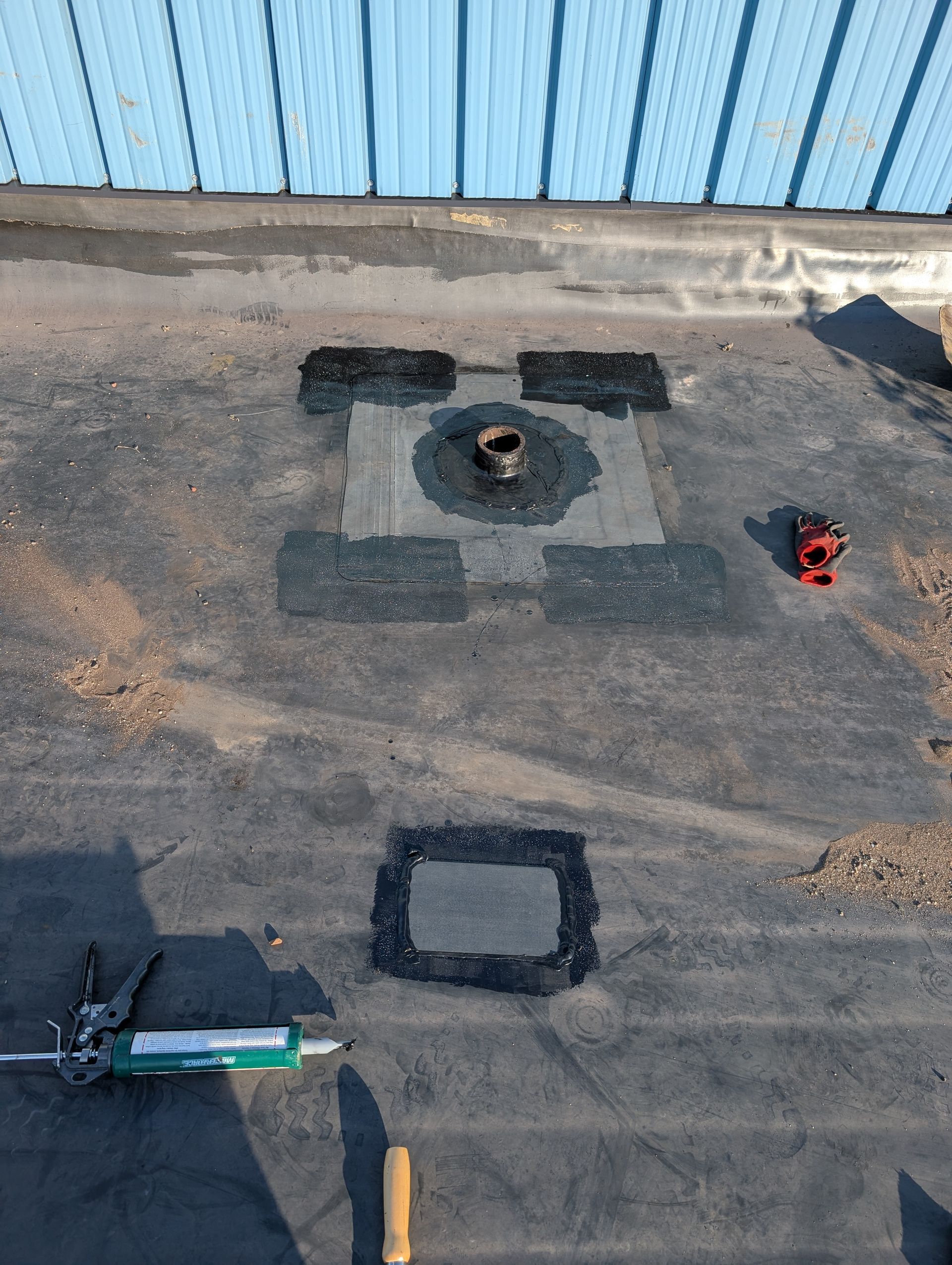 Black flat roof with repair patches around a vent and tools in view.