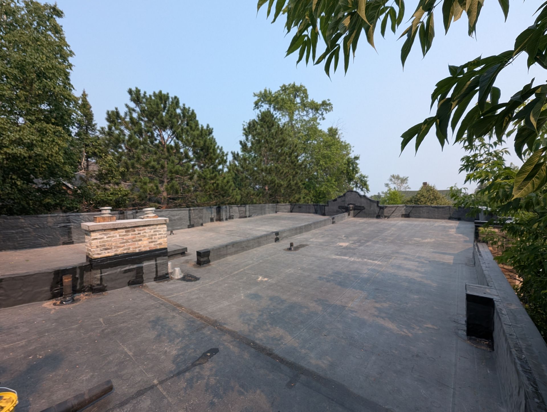 Flat roof with dark covering, surrounded by trees under a clear sky.
