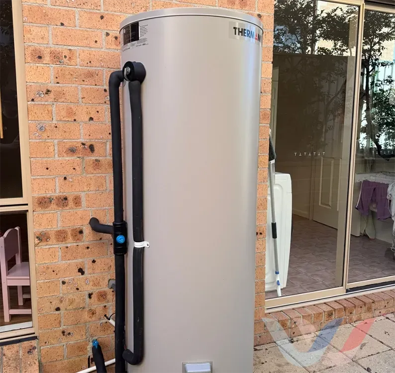 Tan water heater next to a brick wall and sliding glass door. Black pipes run along the side. — Illawater Hot Water Services in Sutherland Shire, NSW