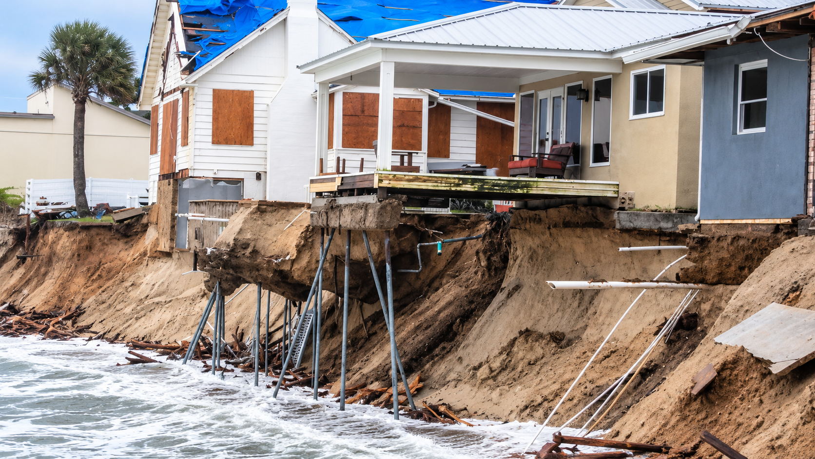Coastal homes perched on eroding cliff above rough ocean waves, with exposed support beams beneath the deck