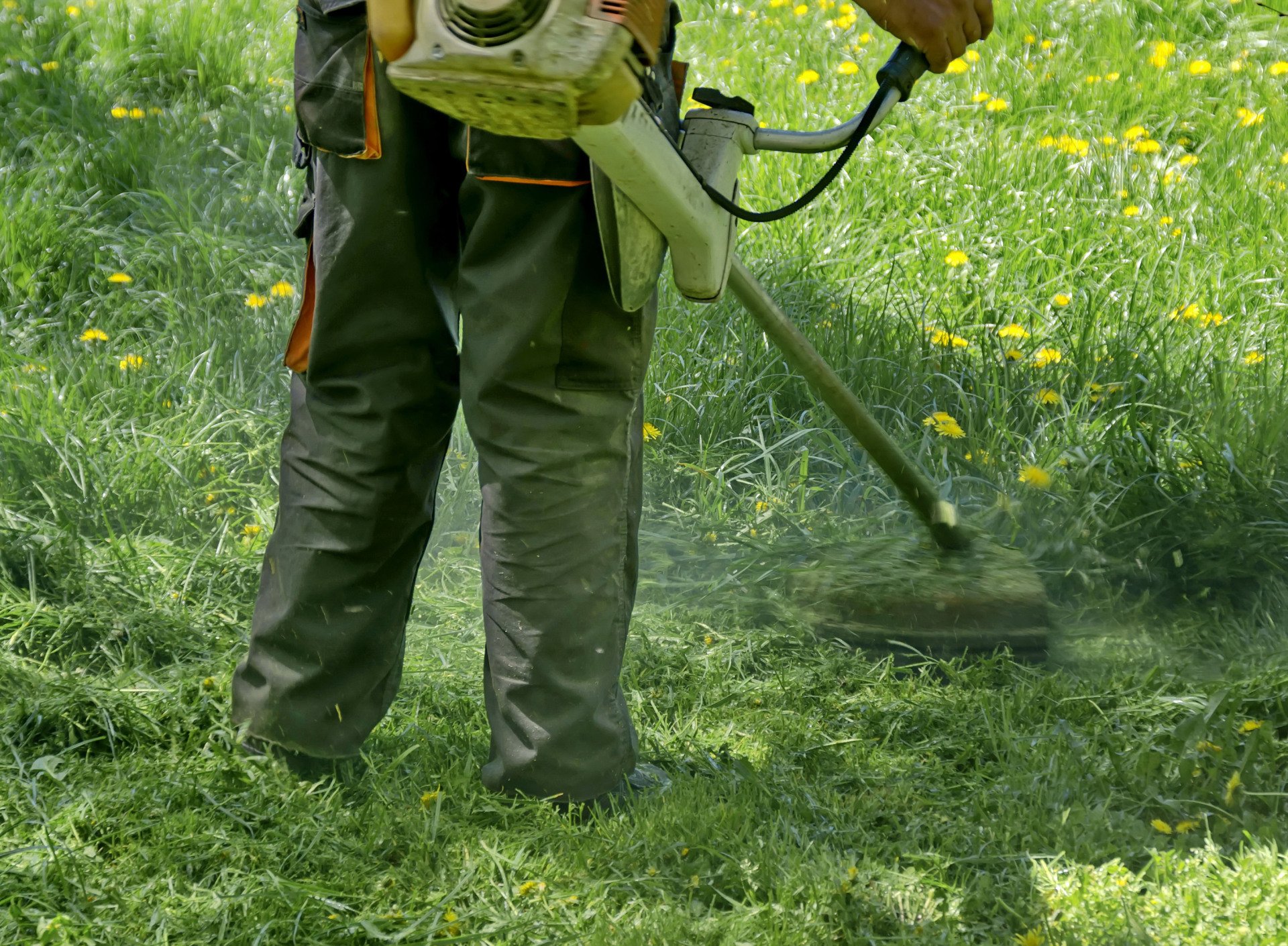 Man trims a hedge with a power trimmer outside a brick house. He wears a yellow shirt.
