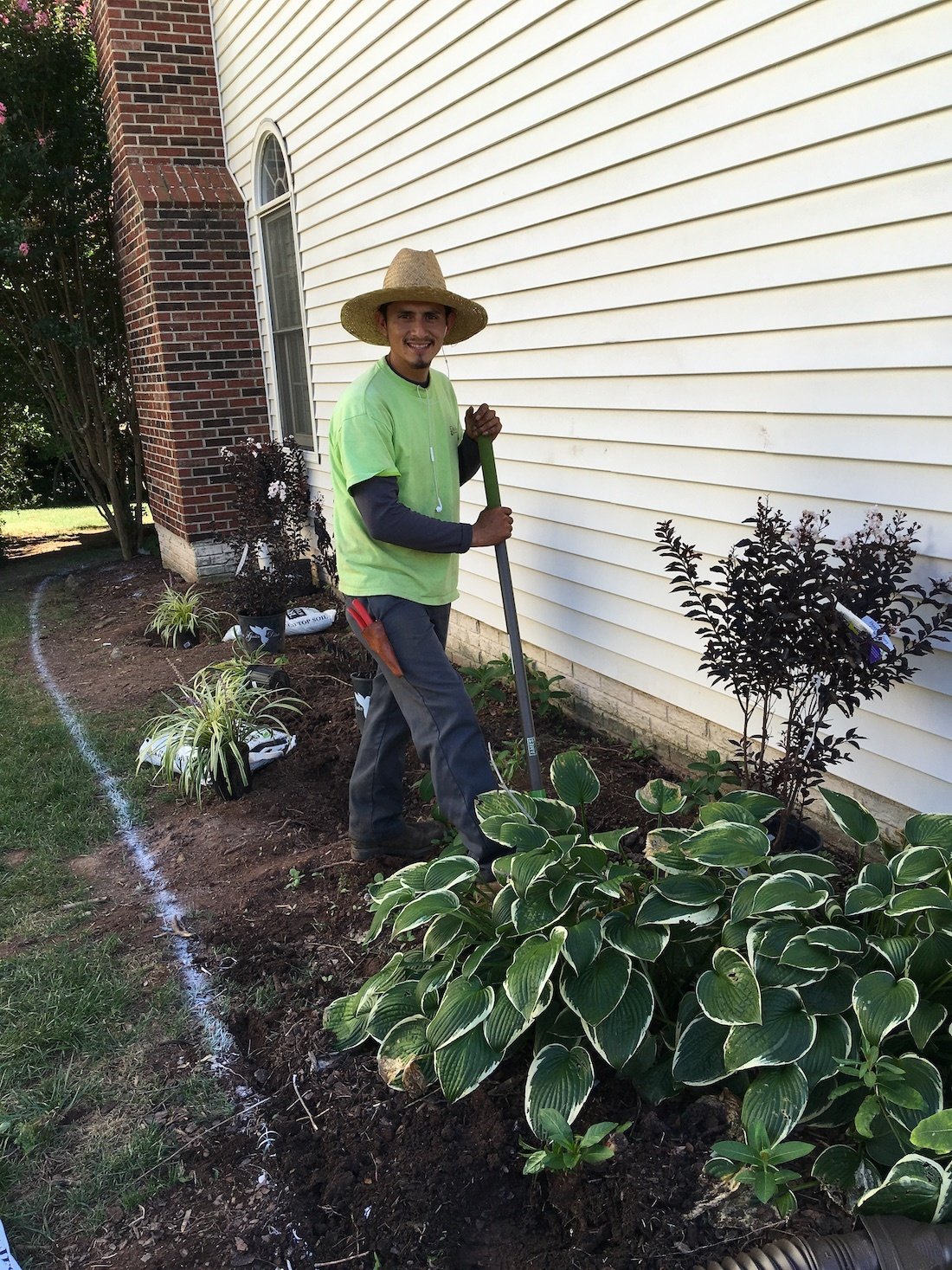 Person in a straw hat and green shirt gardening next to a house with white siding.