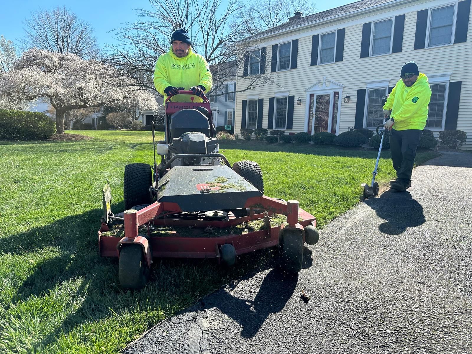 Two landscapers in bright yellow jackets working on a residential lawn, one driving a mower and one using an edger.