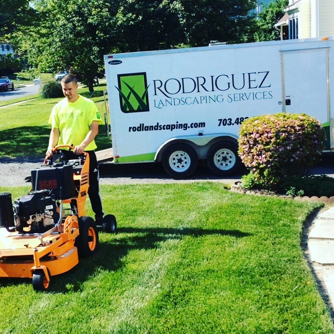 A landscaper in a bright yellow shirt operates a large orange stand-on mower next to a trailer with branded signage.