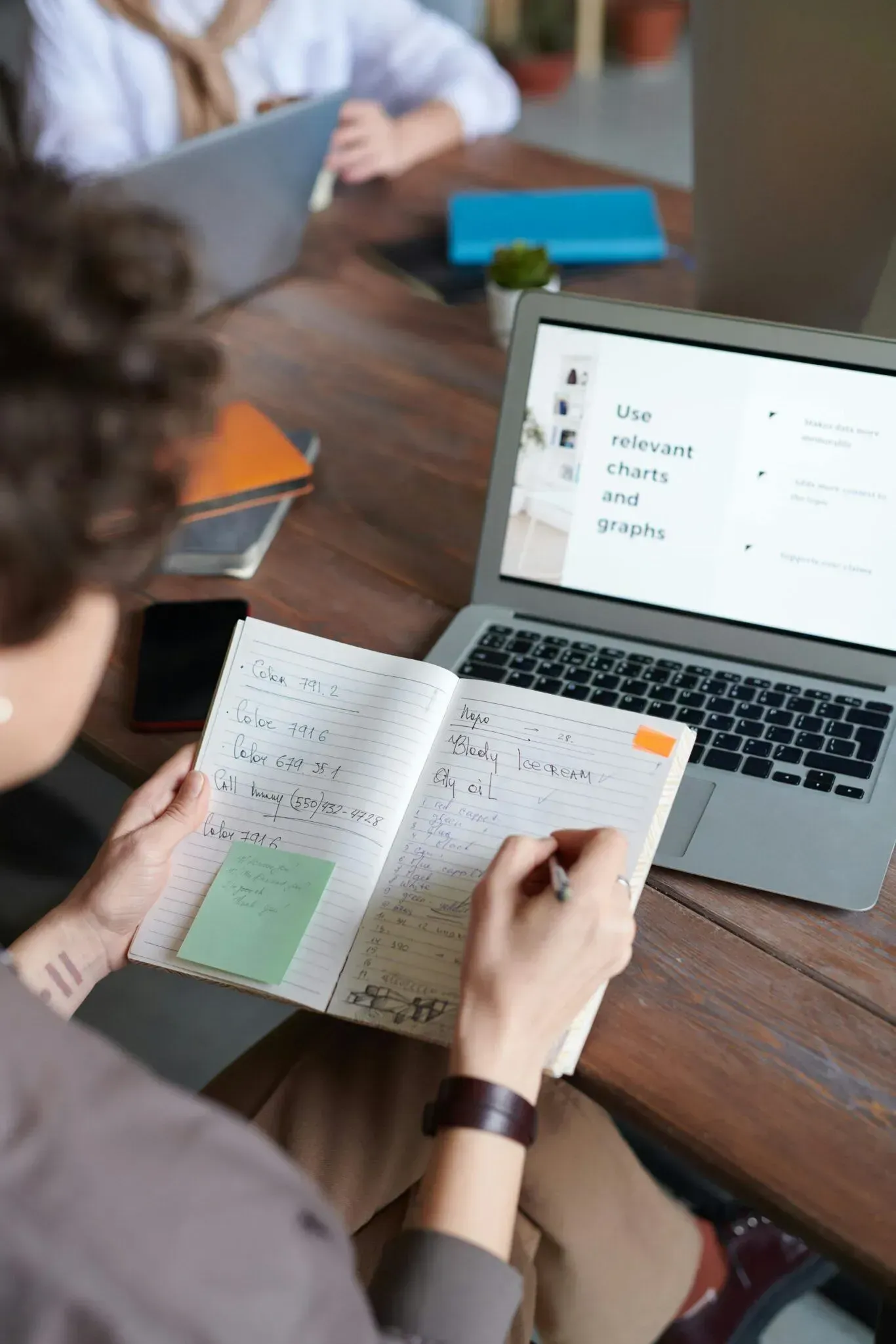 Person writing in a notebook beside a laptop during a meeting at a wooden table.