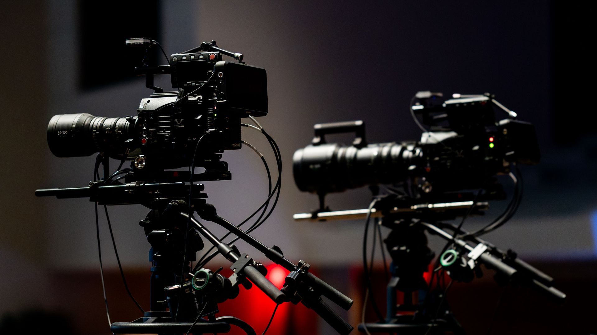 Two black video cameras on tripods in a dim studio with red backlighting