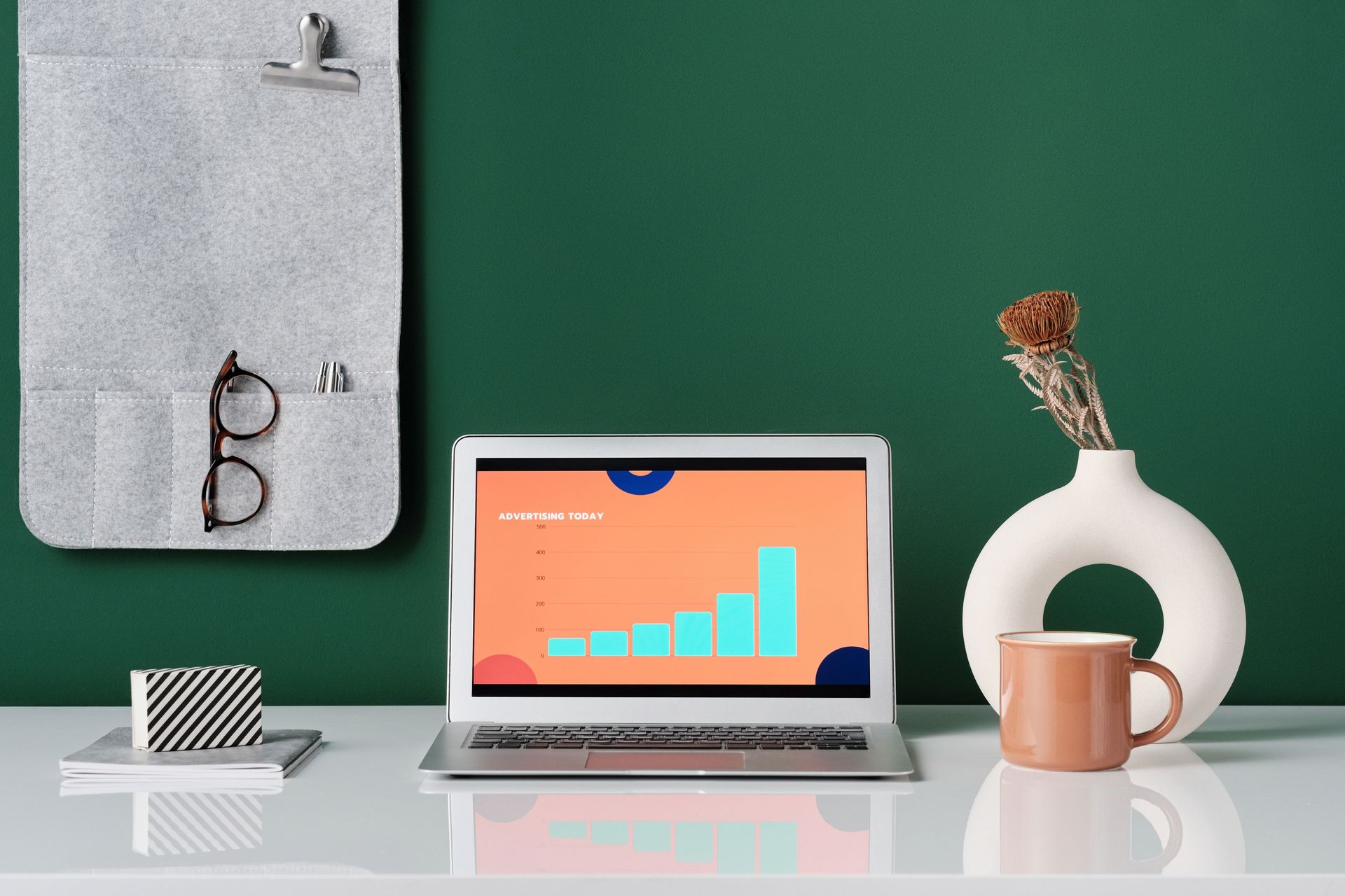 Minimal desk setup with laptop showing a chart, coffee mug, vase, clipboard, and glasses on a green wall background
