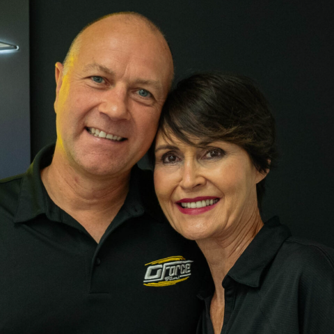 Two smiling people in black shirts posing indoors against a dark background.