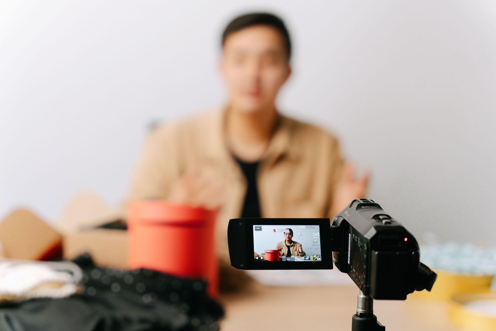 Camera filming a person speaking at a table, with a red cup and clothing in the foreground.