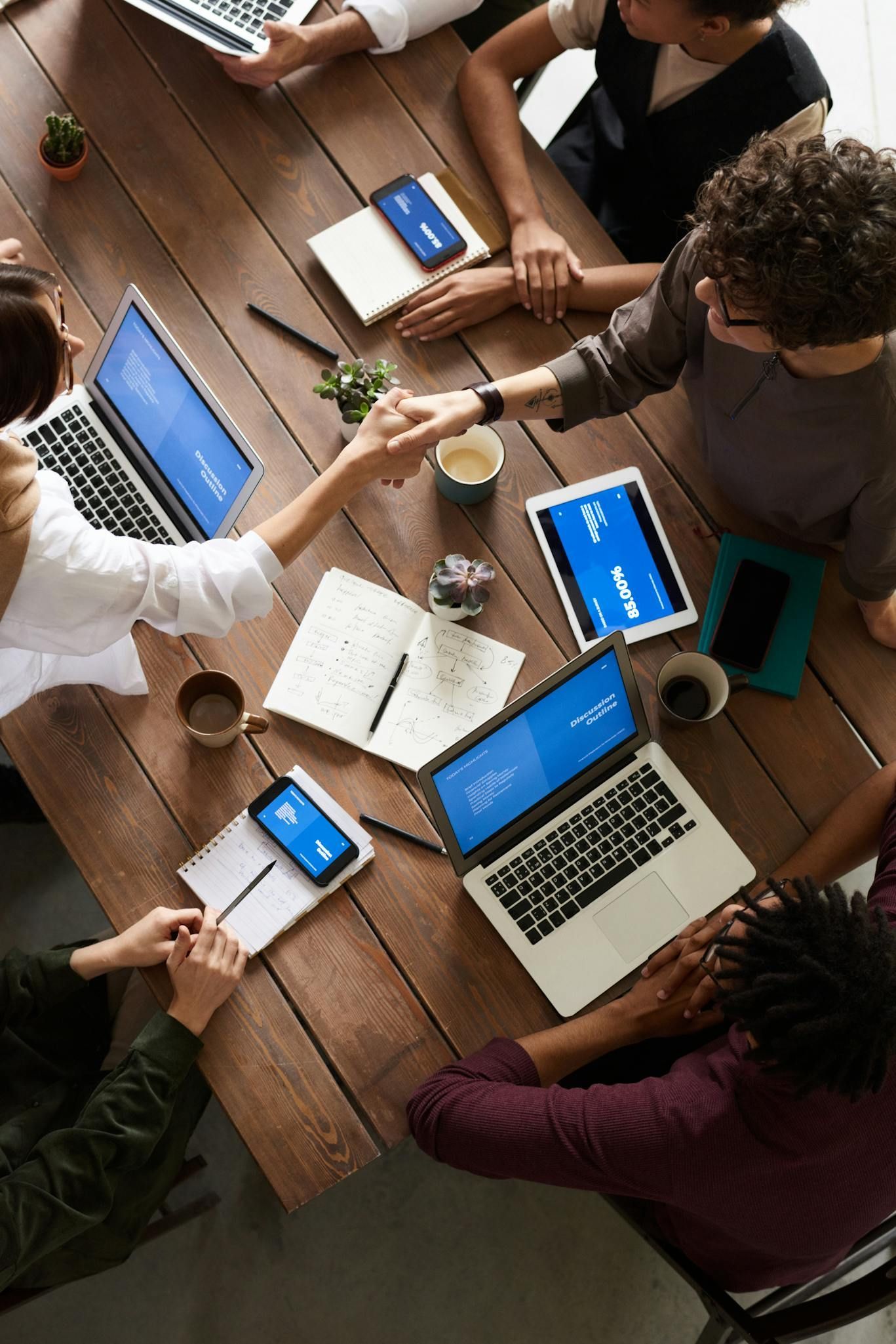 Team meeting around a wooden table with laptops, notebooks, coffee cups, and phones showing blue screens
