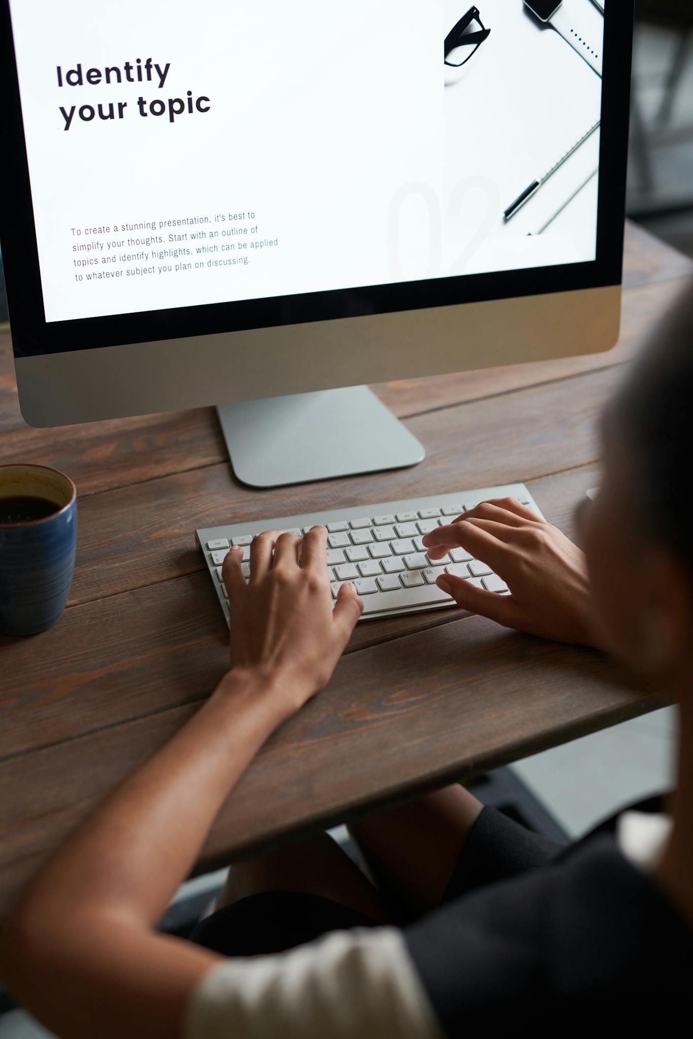Person typing at a desk beside a computer displaying a presentation slide titled “Identify your topic.”