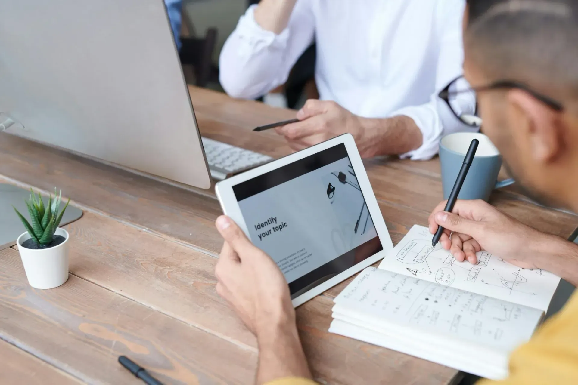 People collaborating at a desk with a tablet, notebook, and laptop in a bright office room