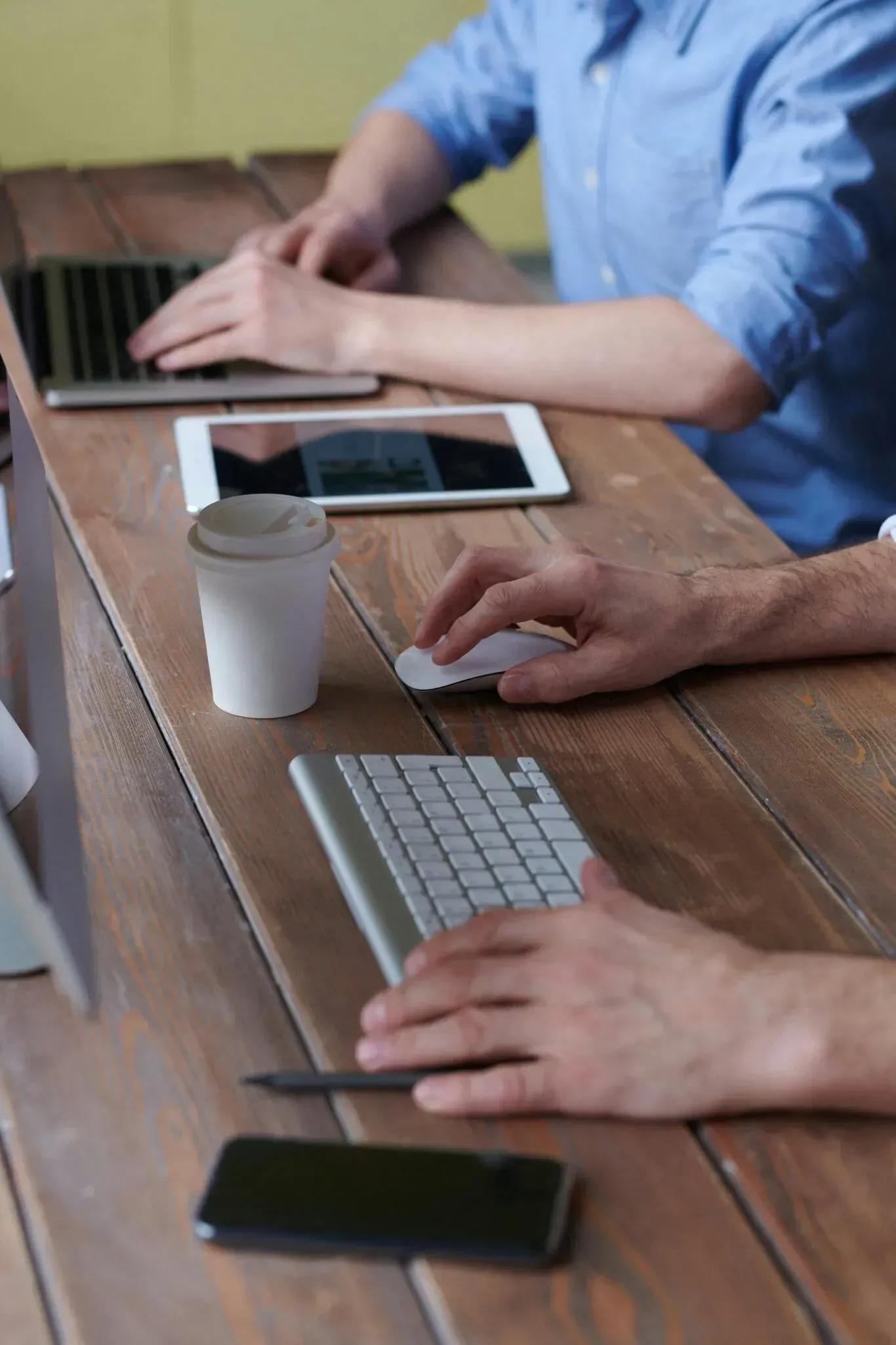 Hands using laptops and keyboards at a wooden table with a tablet, coffee cup, and phone