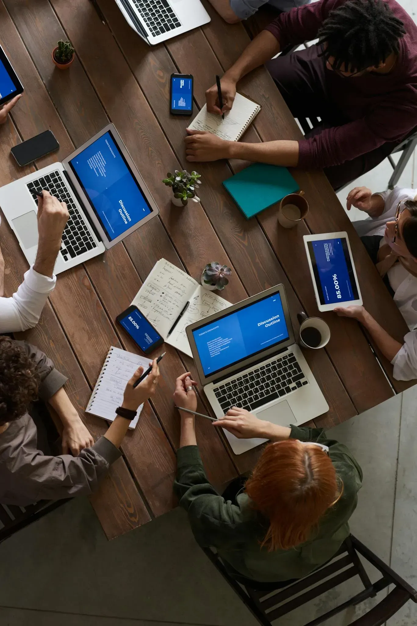 People collaborating around a wooden table with laptops, tablets, notebooks, and coffee.