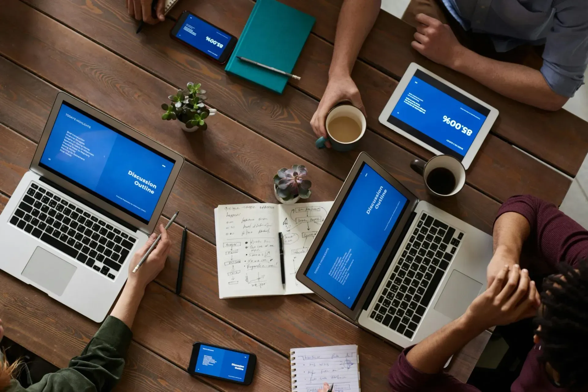 Overhead view of people working at a table with laptops, notebooks, coffee, and a smartphone